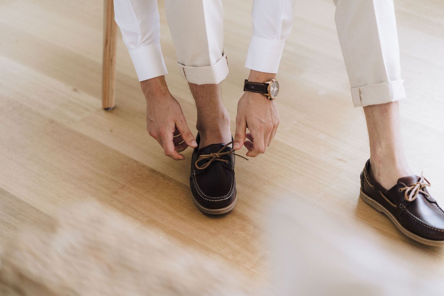 Person tying laces on dark boat shoes while wearing white pants on light wooden floor.