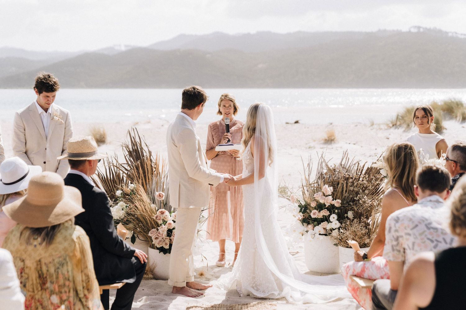 Intimate beachside wedding ceremony with dried pampas grass decorations and guests in sun hats.
