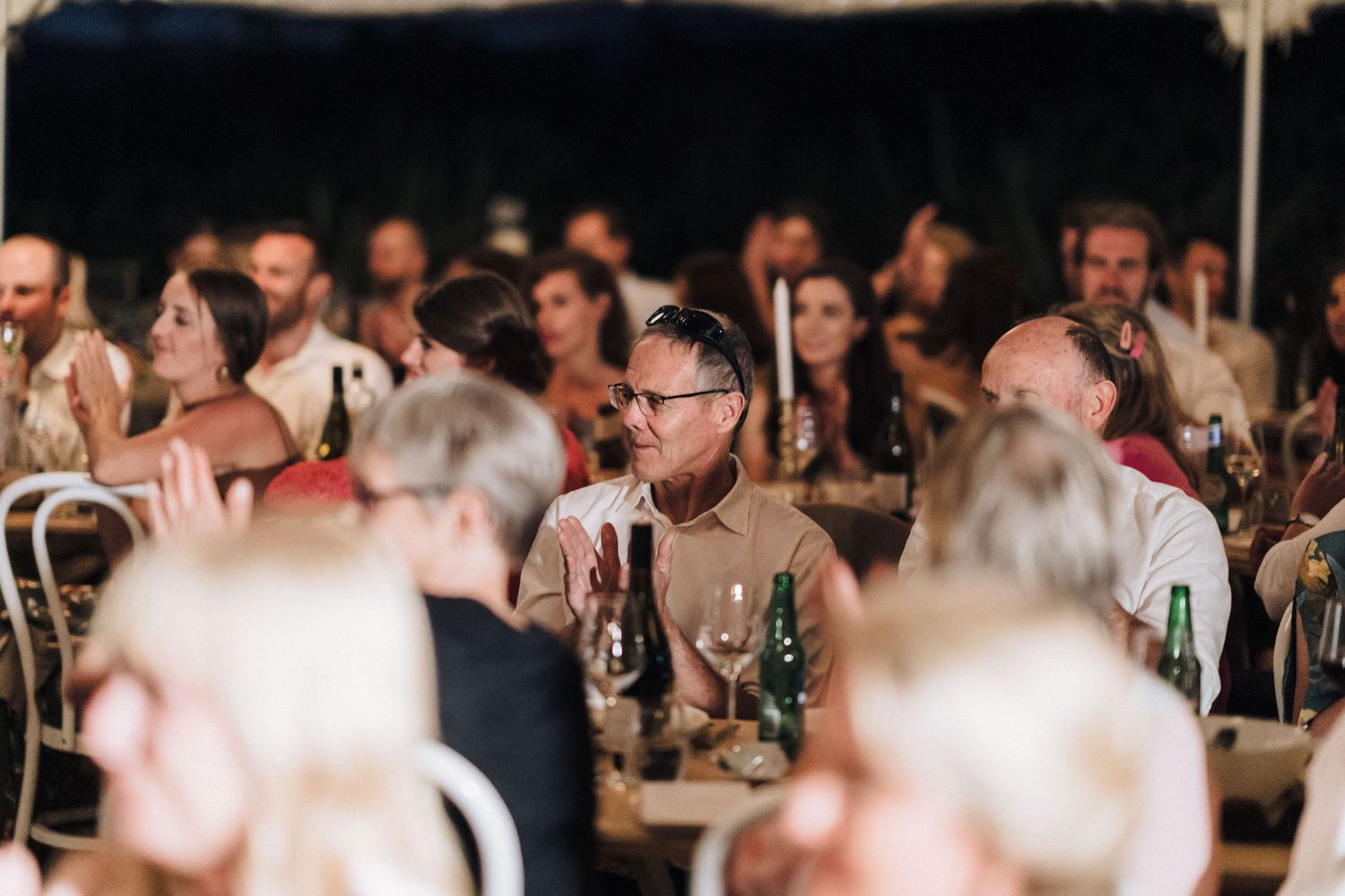 Wedding guests listen attentively during reception under tent lighting.