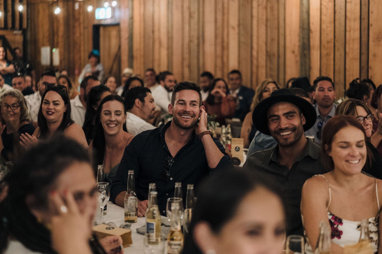 Guests at an indoor reception enjoy themselves with drinks and laughter at tables in a warm wood-paneled venue.