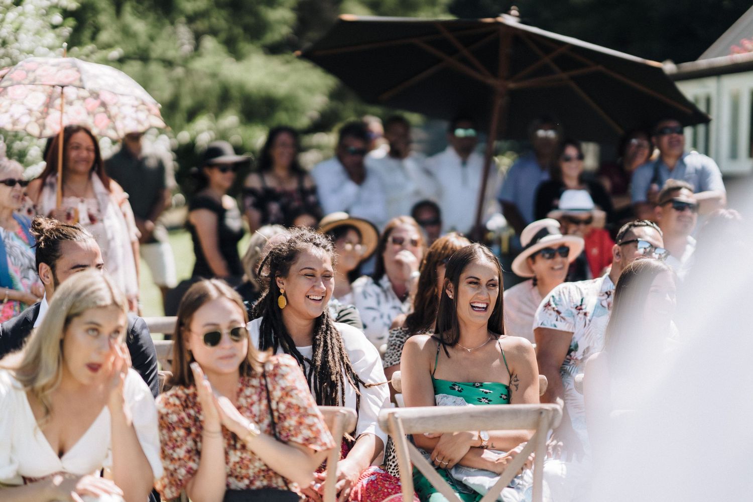 Wedding guests in summer attire enjoy an outdoor celebration under umbrellas and sunglasses.