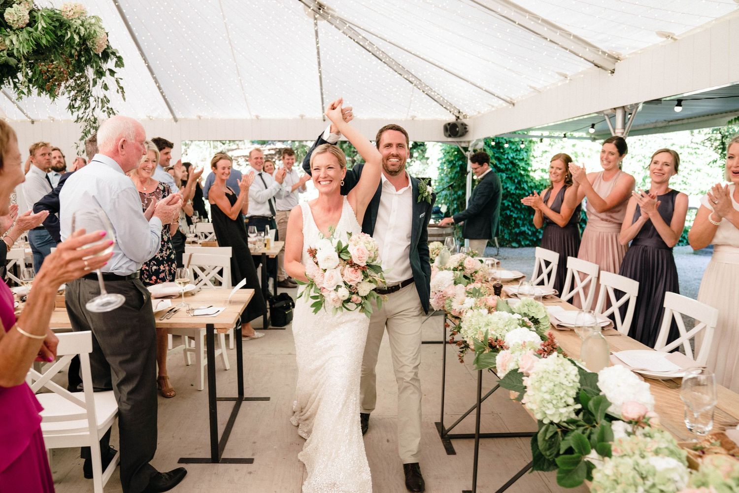 Wedding reception celebration under white tent with guests applauding couple.