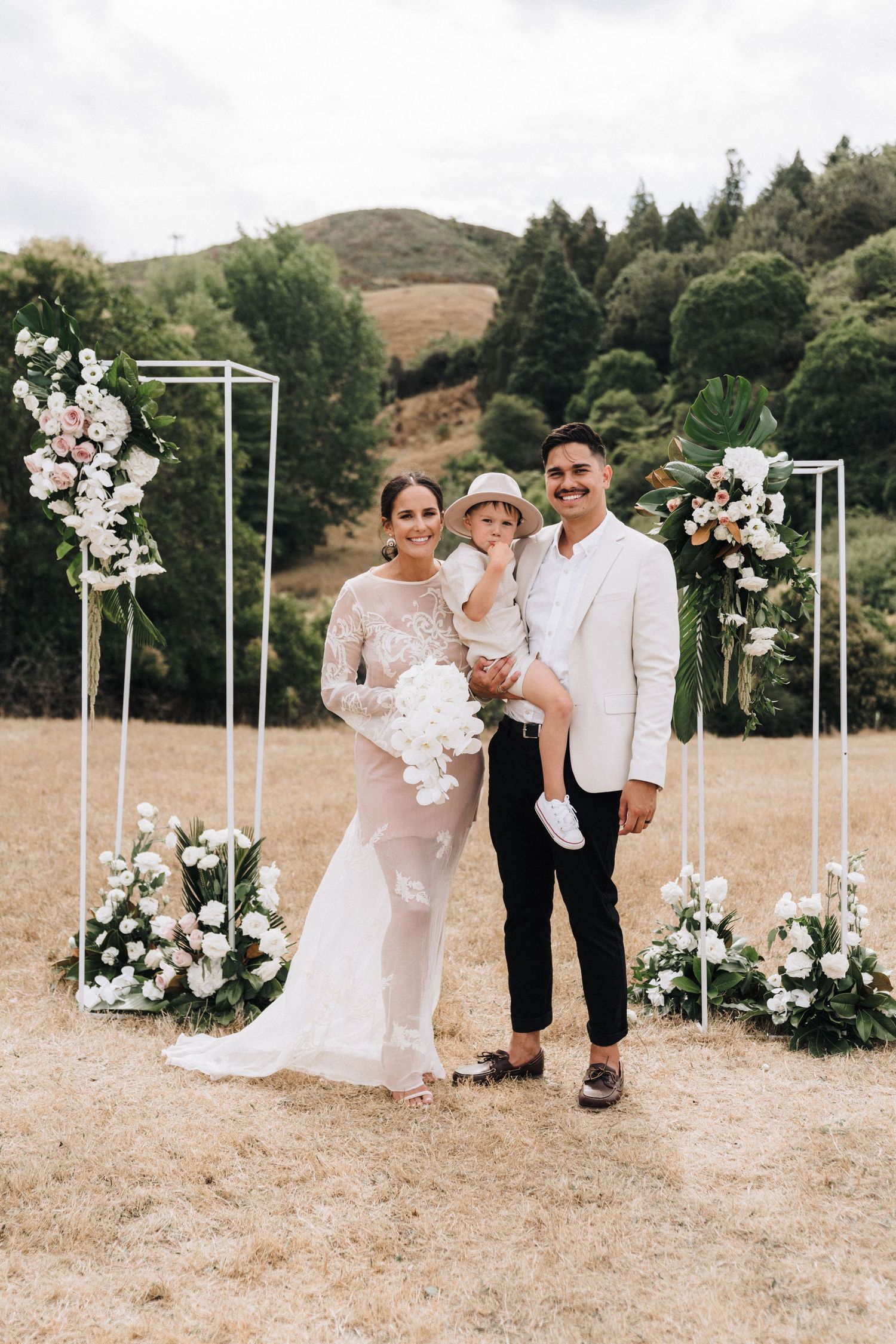 Wedding couple poses with child under elegant white floral arches against rolling hills and forest backdrop.