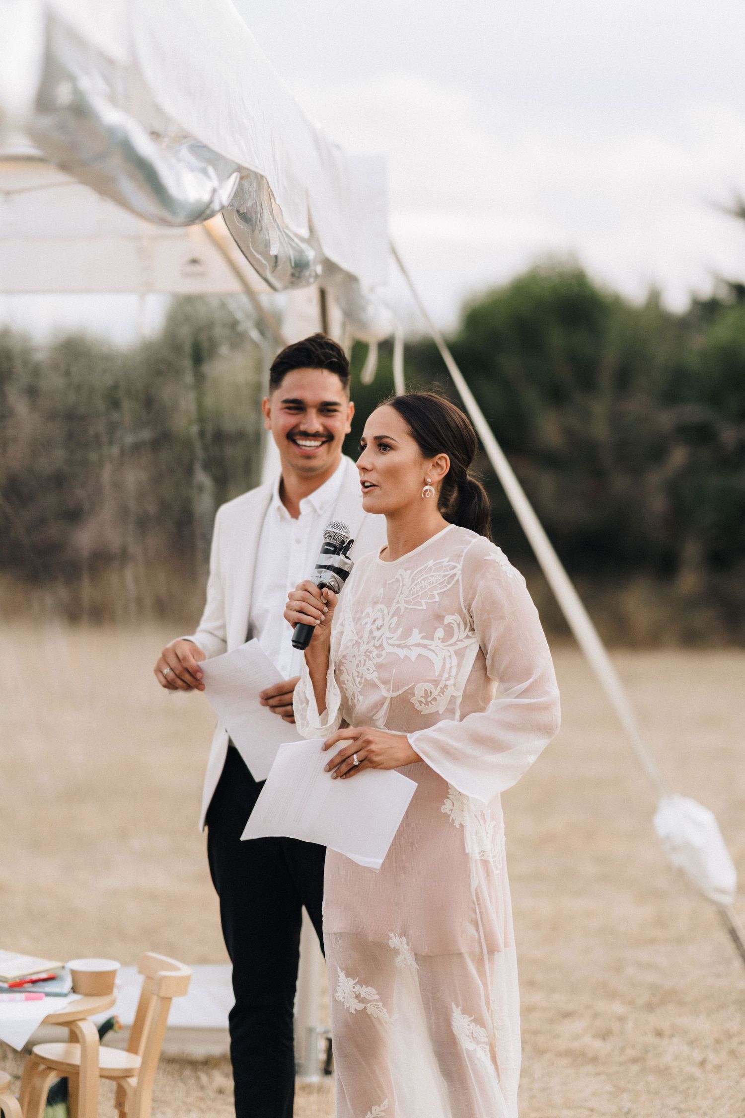 A couple shares a happy moment during wedding speeches under white banners in an outdoor setting.