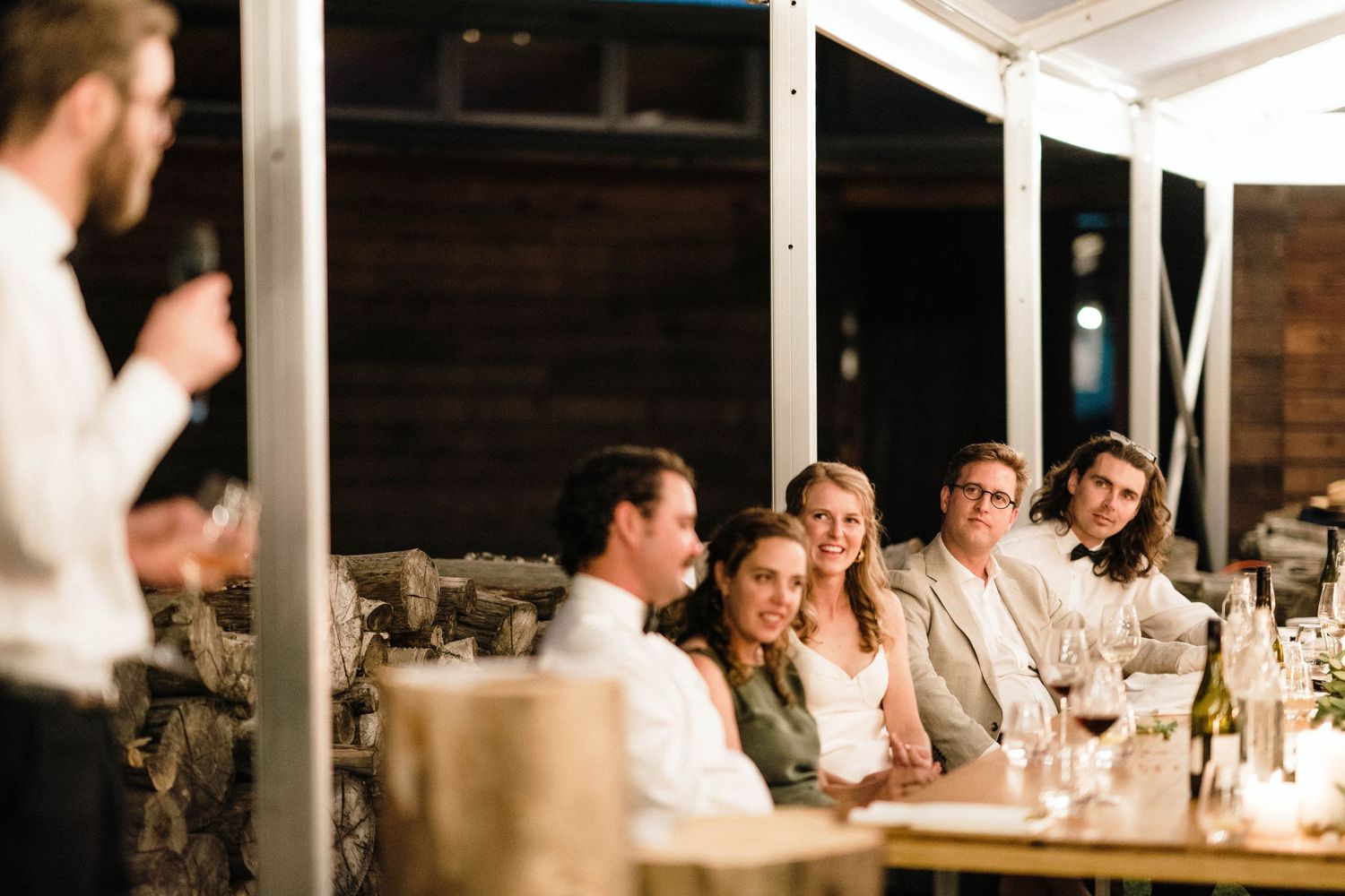 Guests seated at tables enjoy wedding speeches during an evening outdoor celebration.