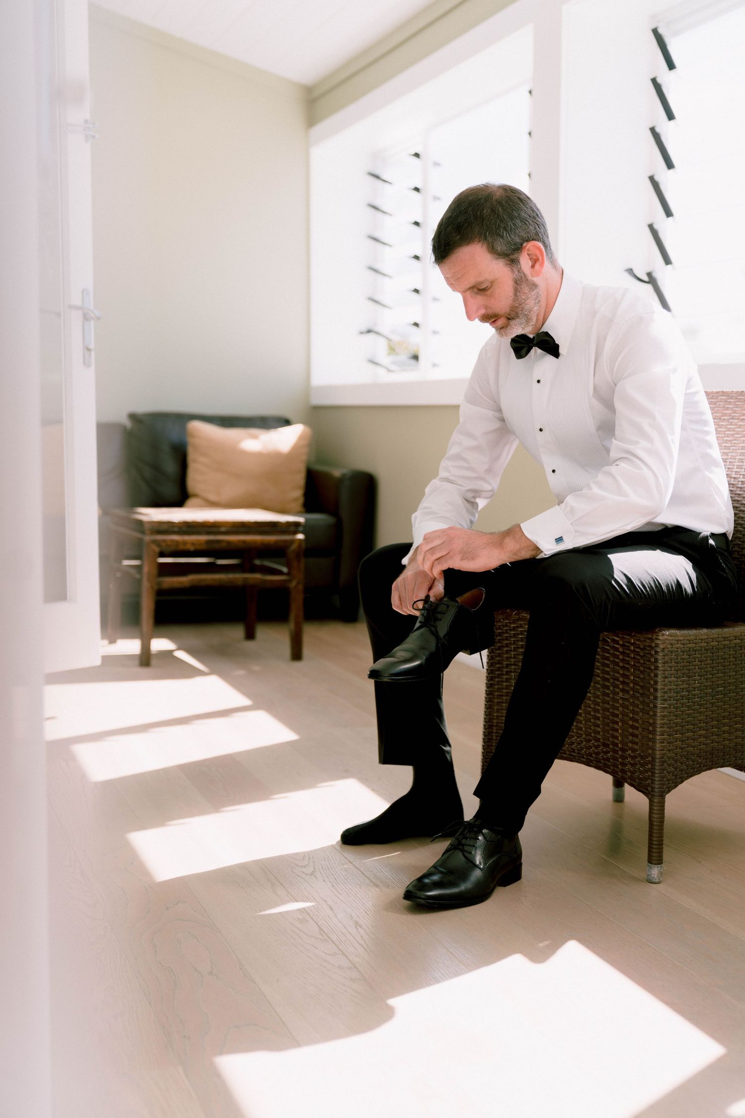 Groom in formal attire adjusts his shoes while sitting in sunlit room with modern decor.