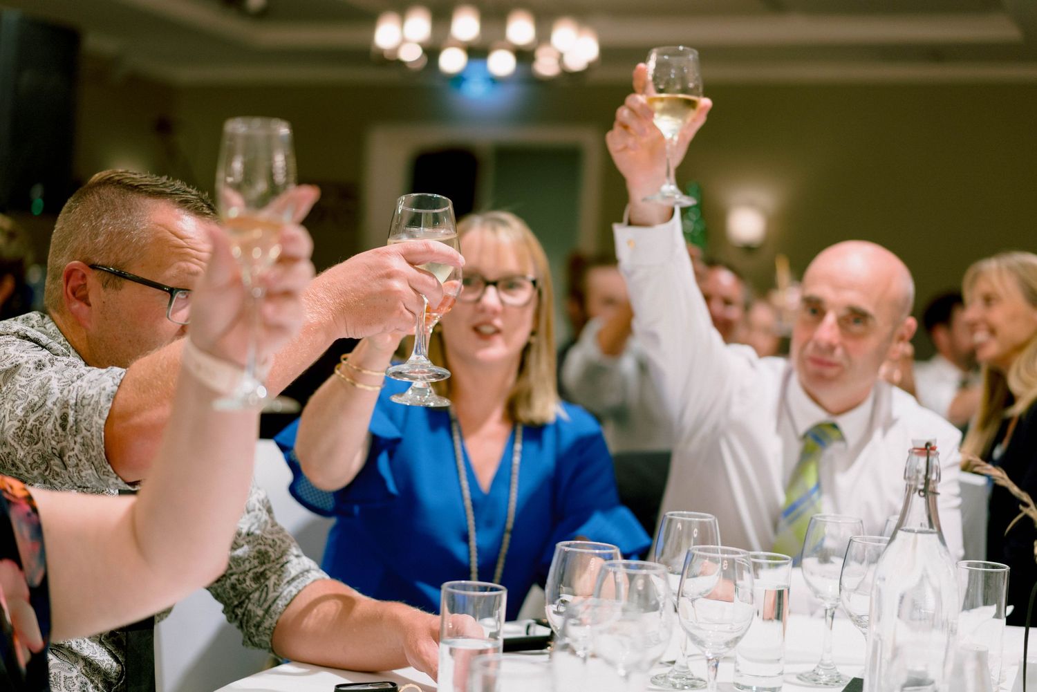 Wedding reception guests seated at tables with glasses raised during evening celebration.