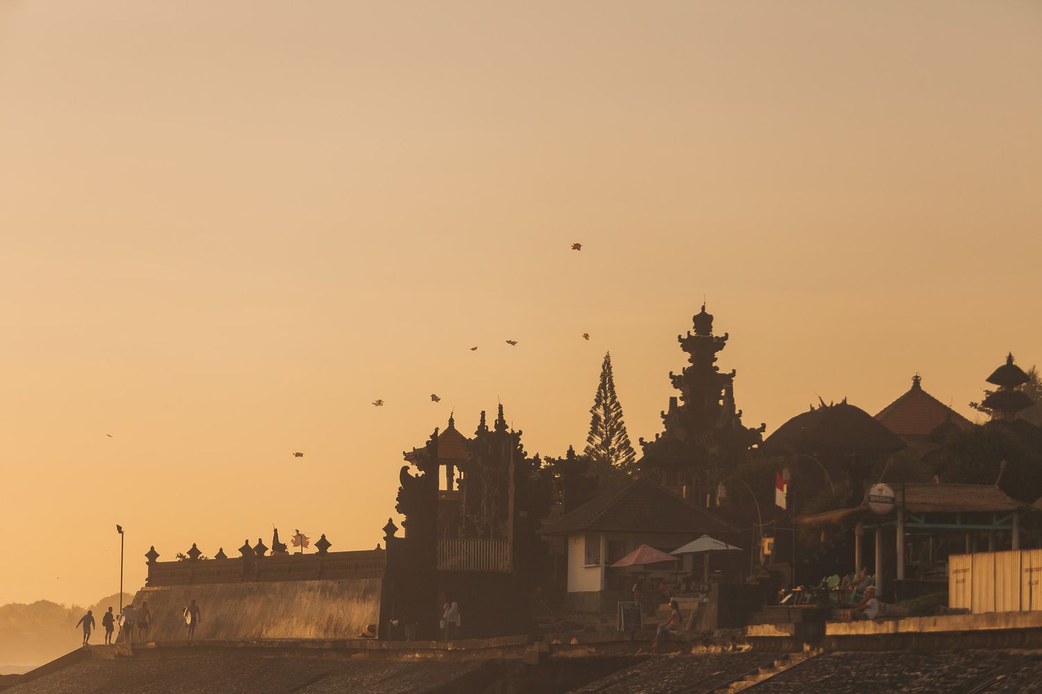 Golden sunset silhouettes ancient temples and buildings along a coastal skyline with birds flying overhead.