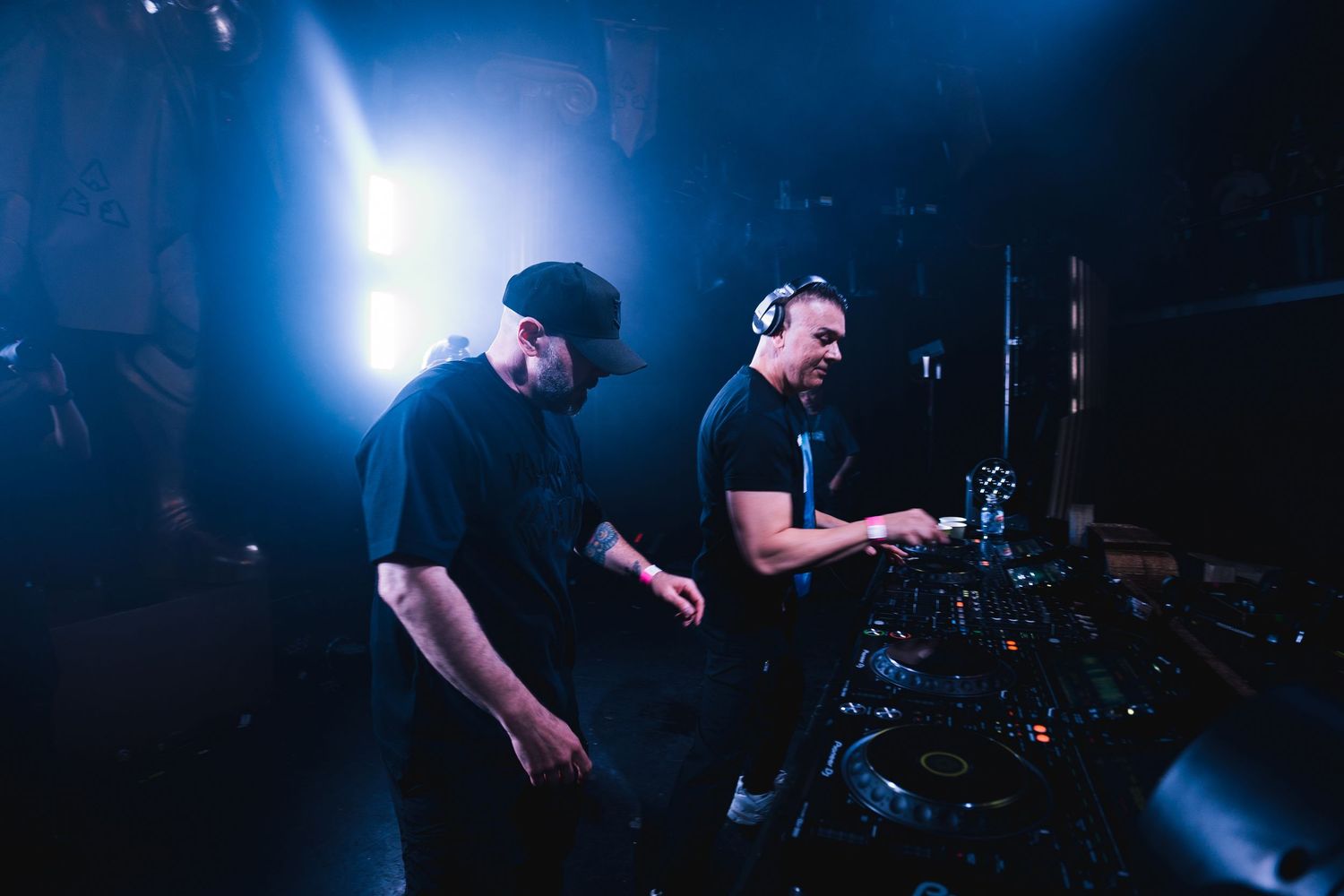 Two DJs perform together on stage at a nightclub with blue lighting and electronic music equipment in a dark environment.
