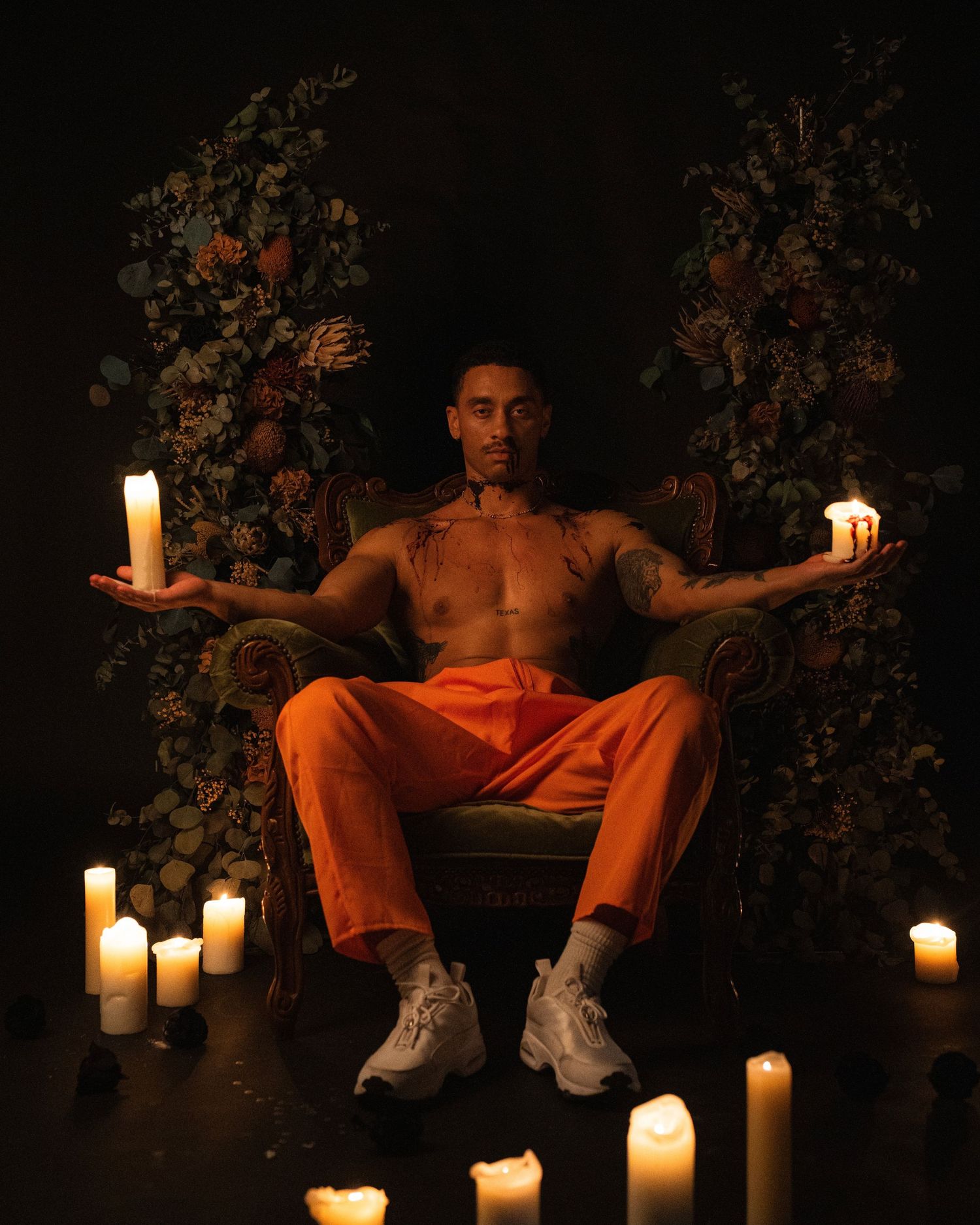 Moody portrait with figure seated on chair surrounded by glowing candles and floral decorations in dark setting.