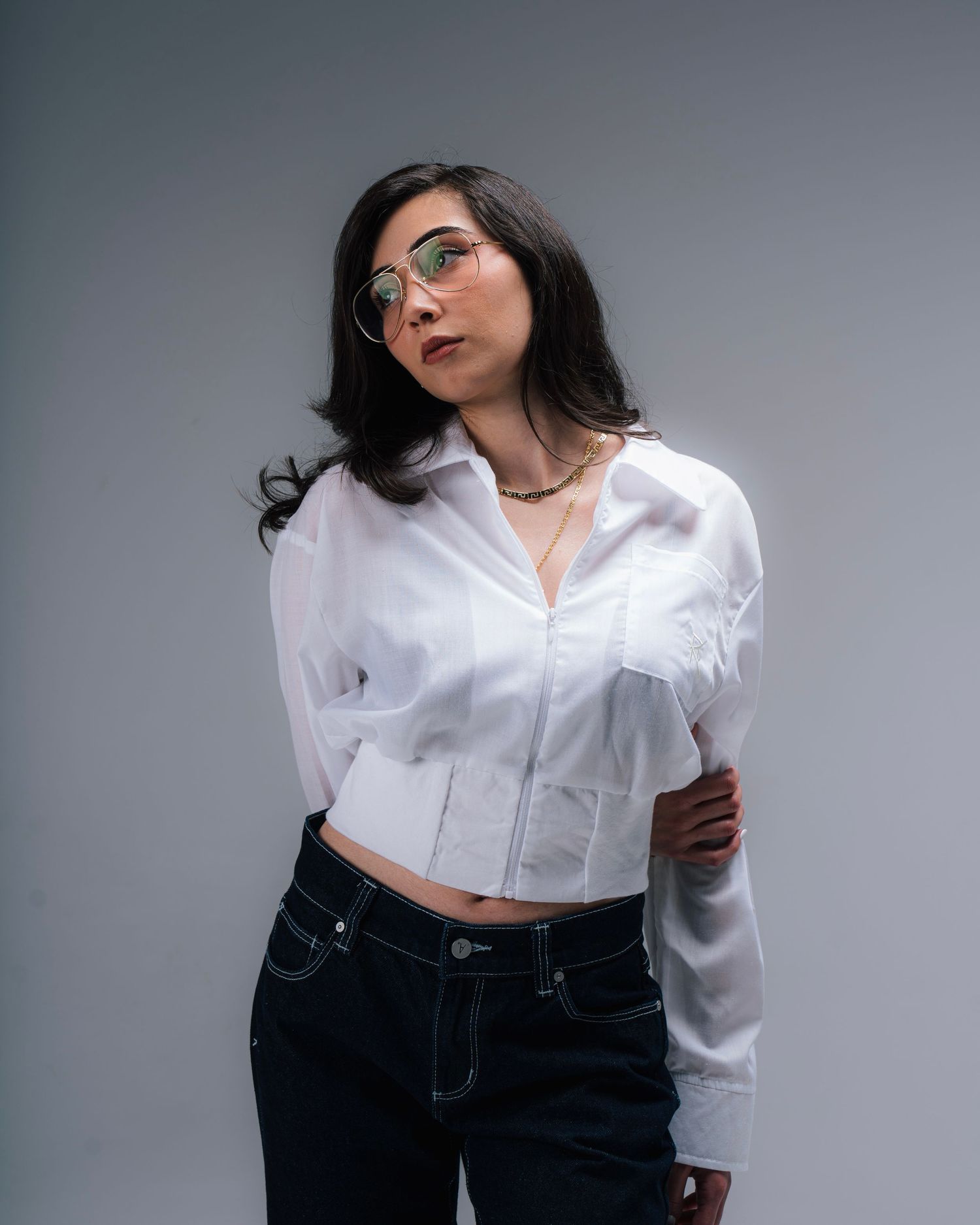 A portrait of someone wearing glasses, a cropped white button-up shirt and dark jeans against a gray background.