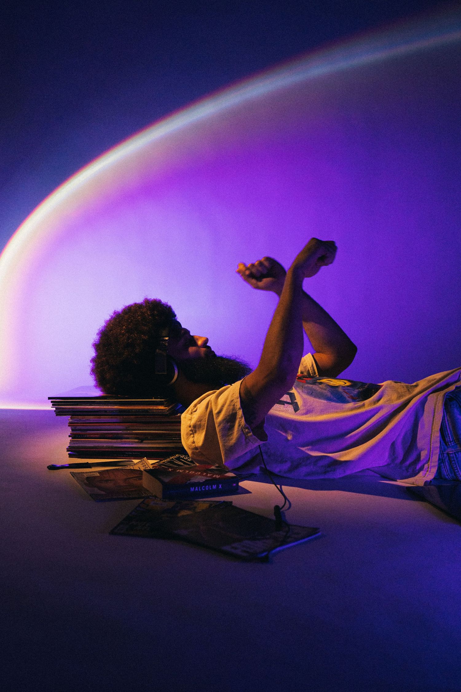 Person lying on stack of vinyl records against purple background in artistic lighting with glowing rainbow arc effect.