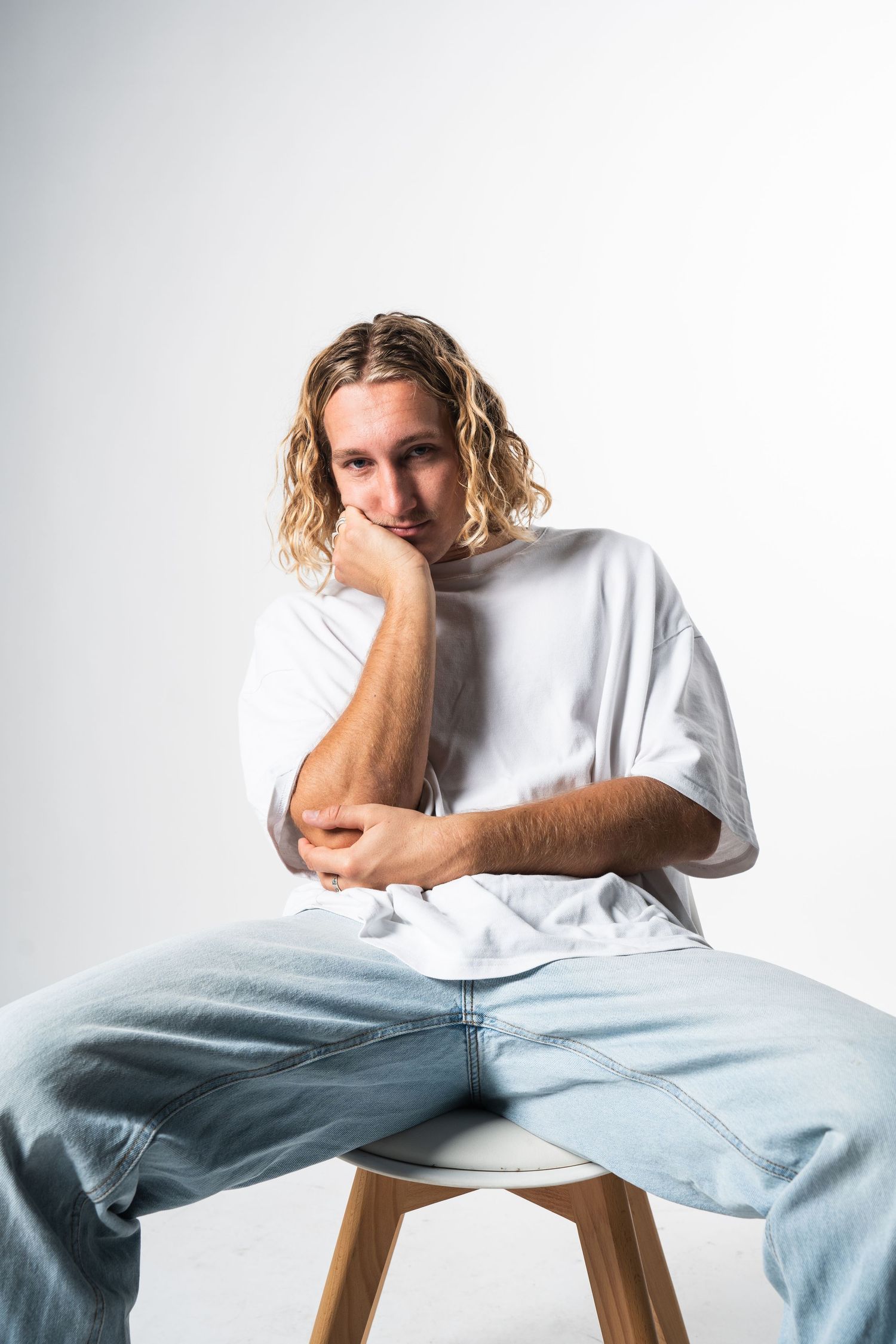 Person wearing white t-shirt and light blue jeans sits casually on wooden stool against white backdrop in three poses.