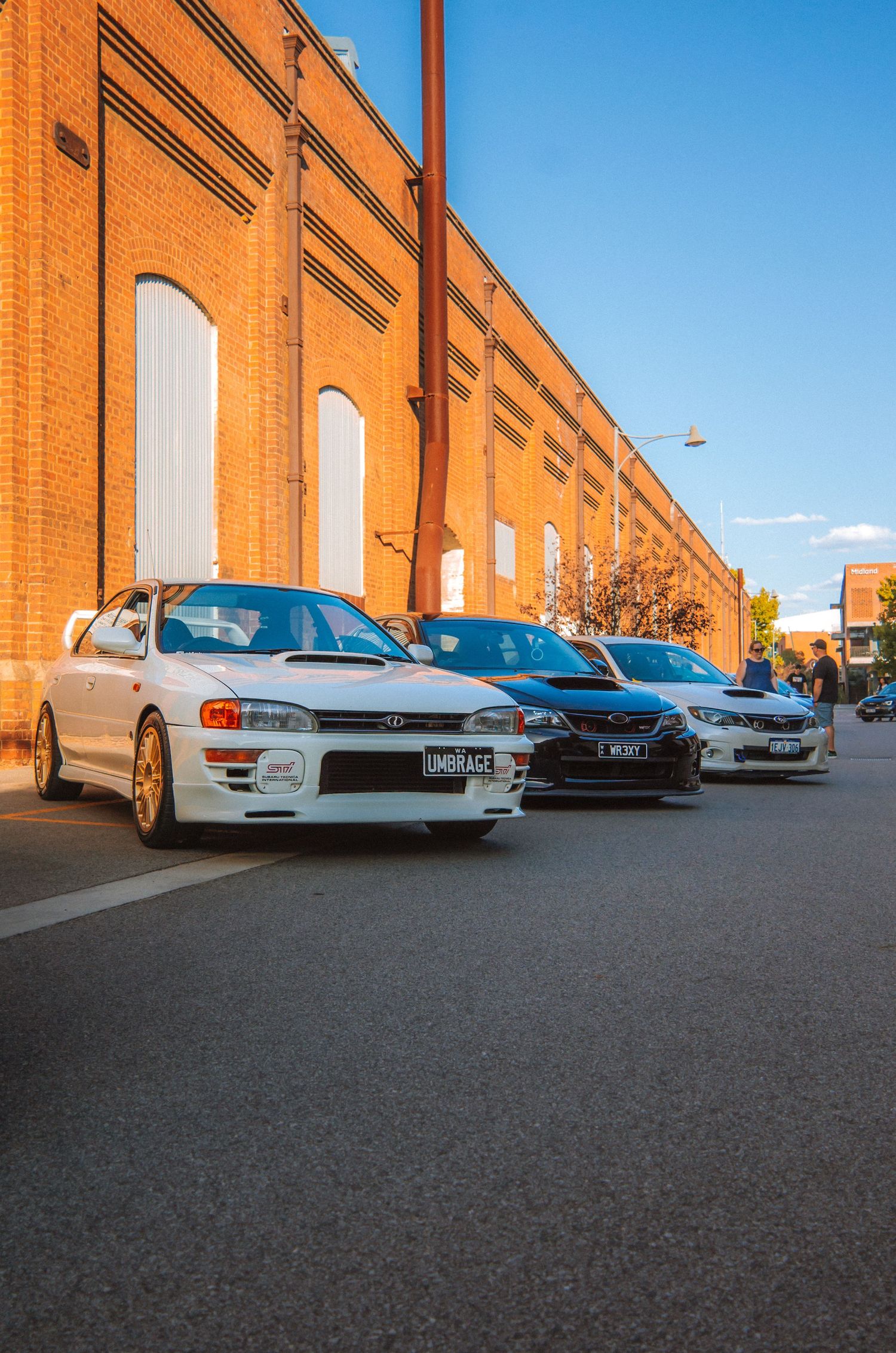 Classic Subaru Impreza and modern sports cars lined up against brick building facade.