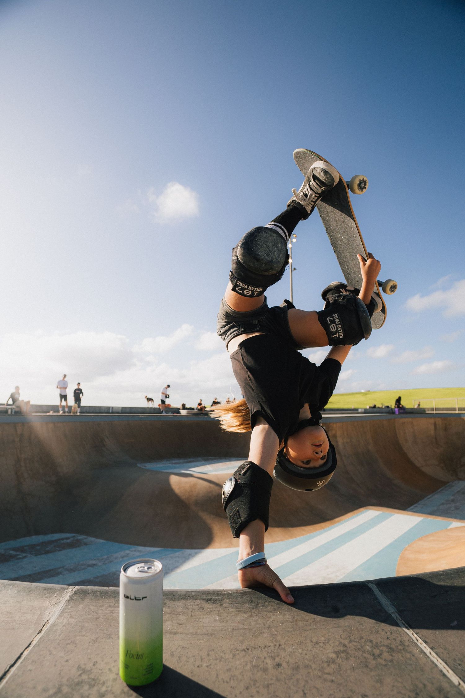 Skateboarder executing inverted aerial maneuver at skate park with green drink container.