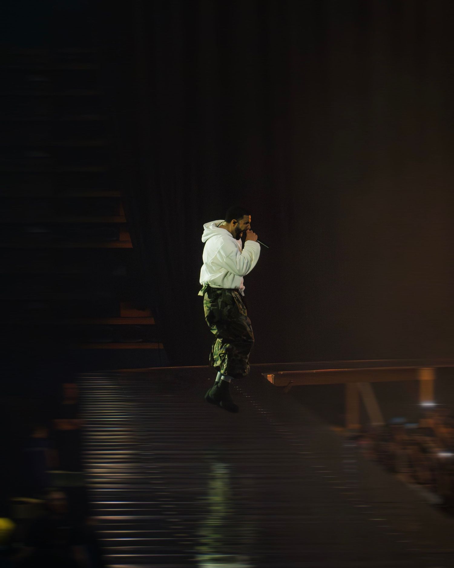 A person in green cargo pants and white top performs on a darkened stage under dramatic lighting.
