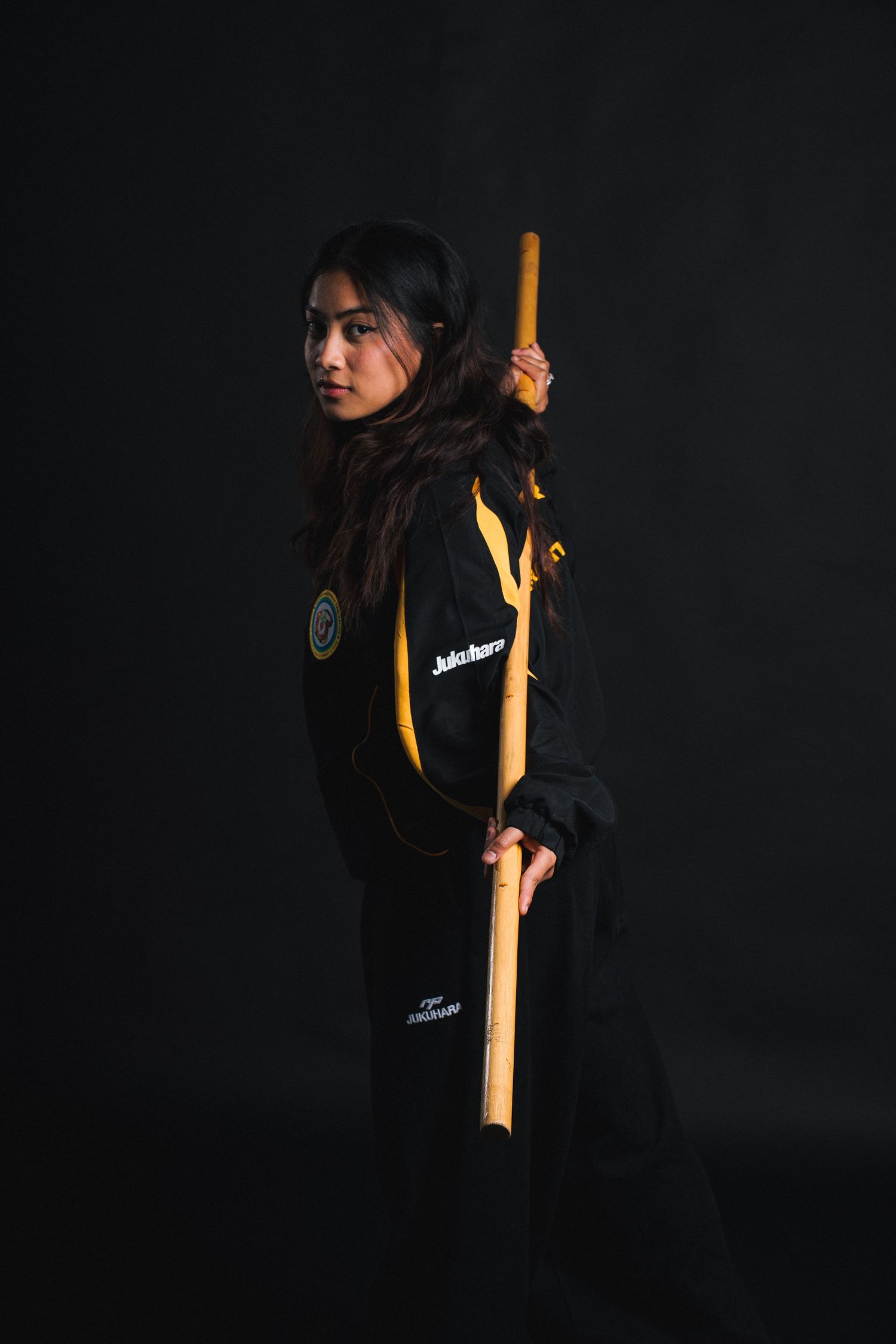 Dramatic portrait of person in black and yellow athletic wear holding wooden staff against dark background.