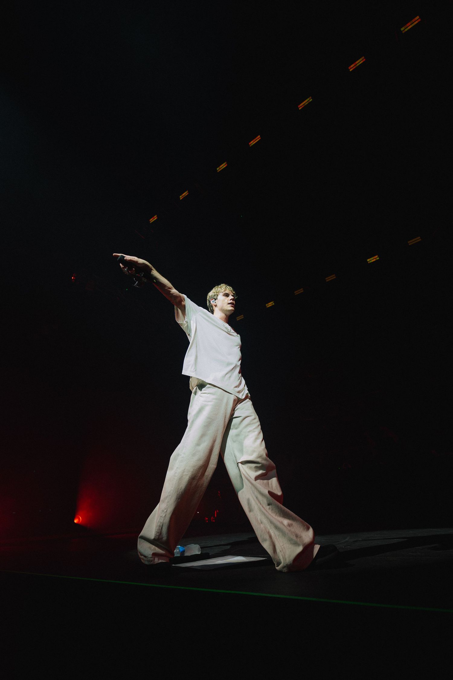 A performer in white clothing strikes a dramatic pose on a dark stage with red lighting and floating orange elements.