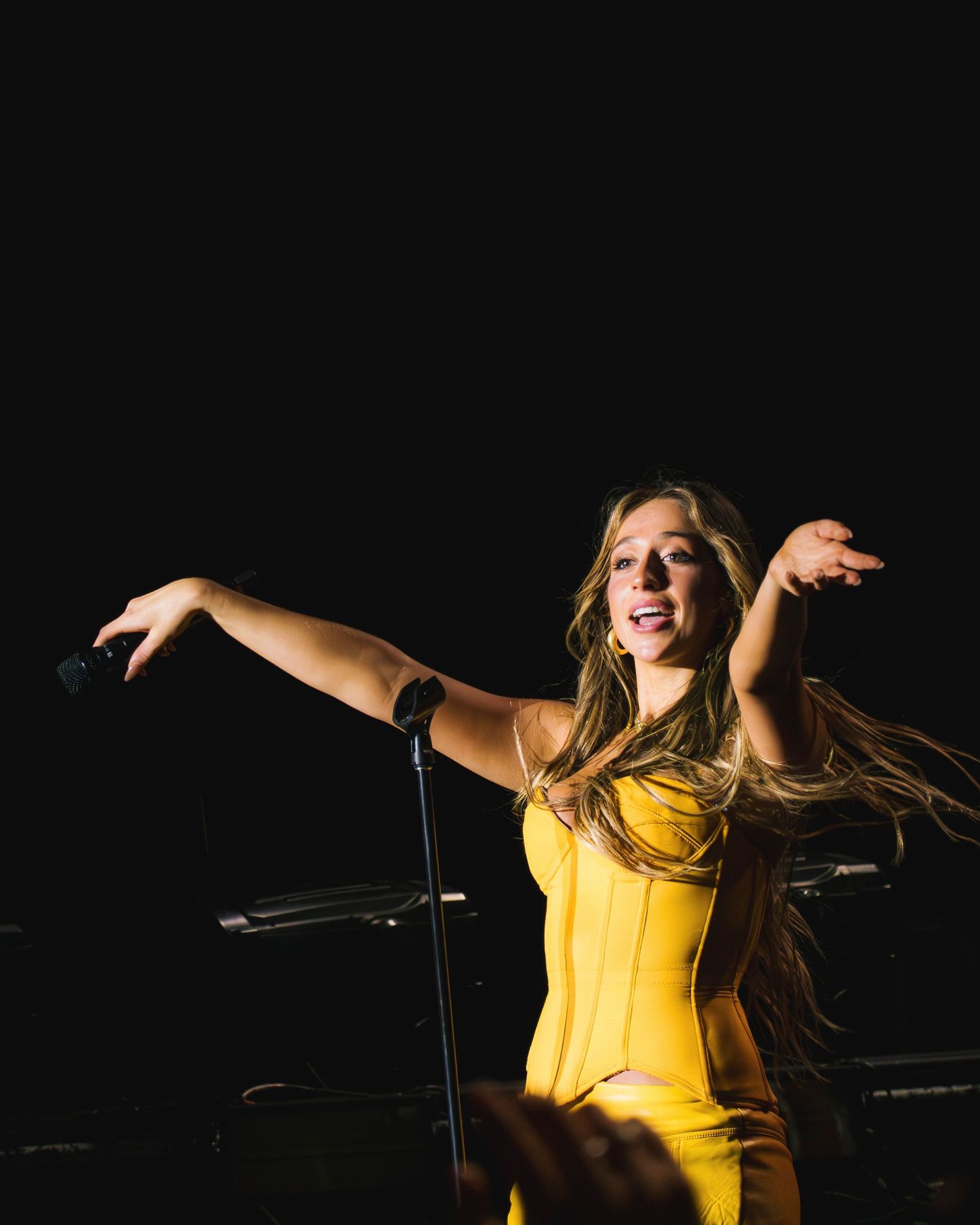 Performer in bright yellow dress dances energetically on dark stage with arms outstretched during live show.