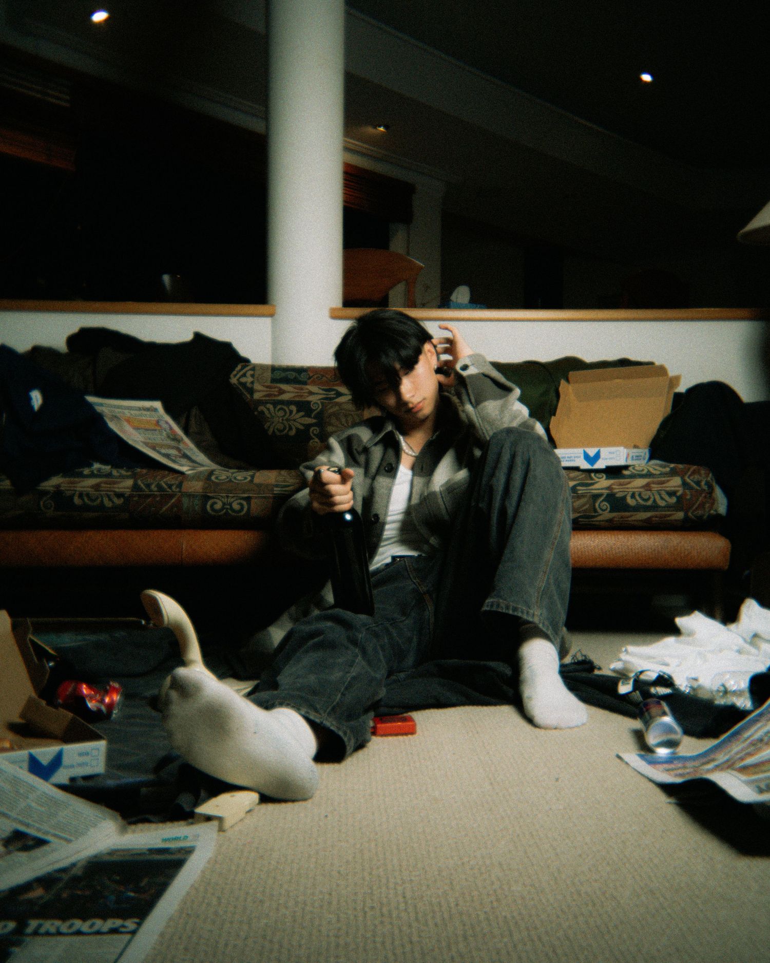 Someone sitting on the floor of a messy bedroom wearing headphones and loose clothing among scattered papers and items.