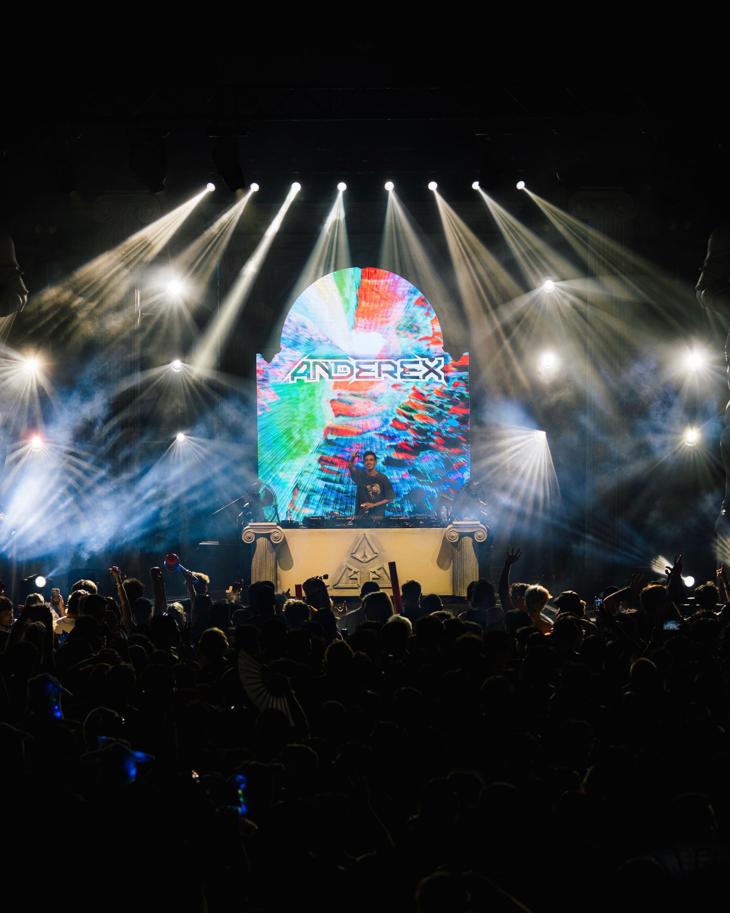 Concert stage with dramatic lighting and a colorful digital display screen illuminated by multiple spotlights over a crowd.