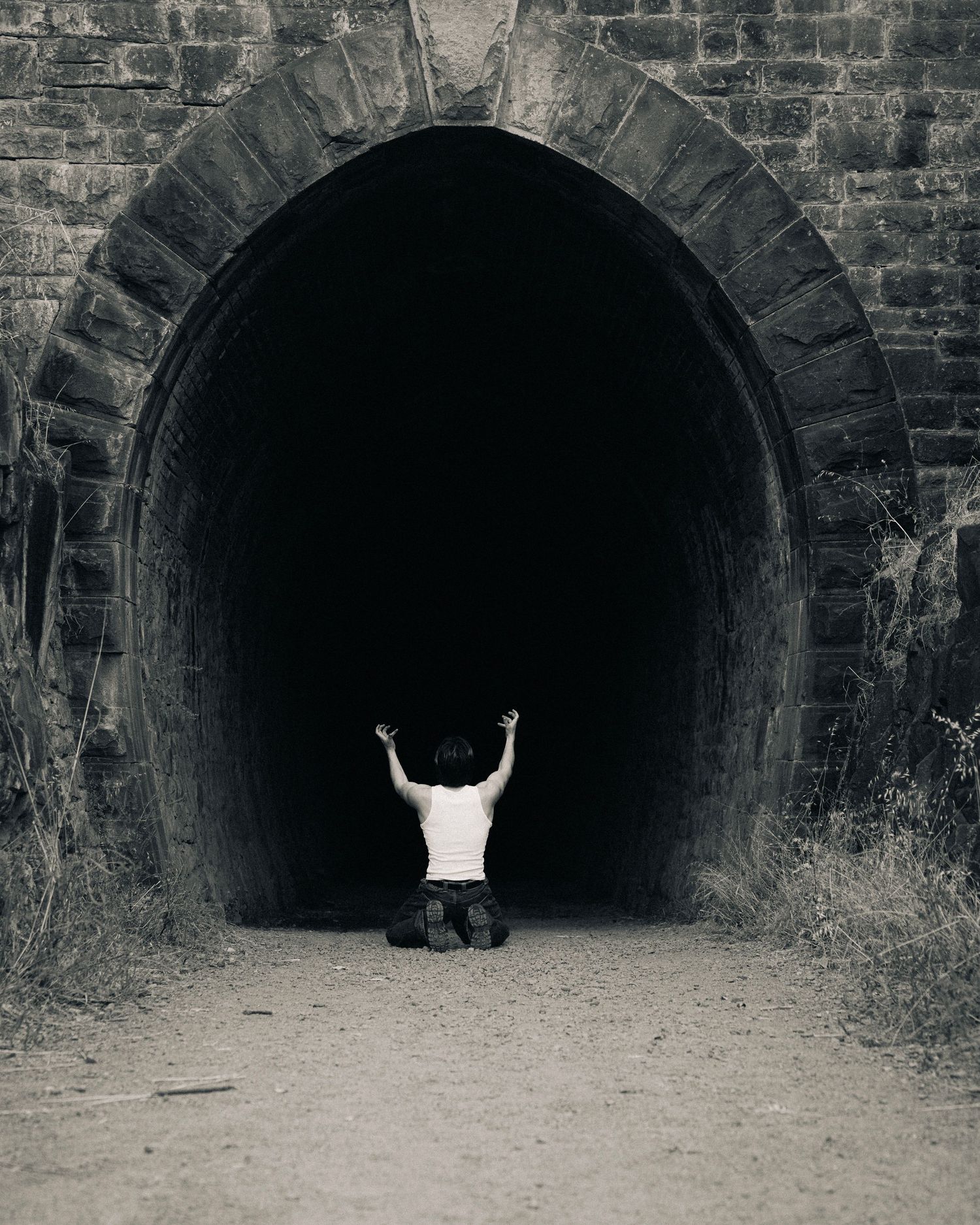 Figure silhouetted in white kneels at entrance of dark stone tunnel with arched opening.