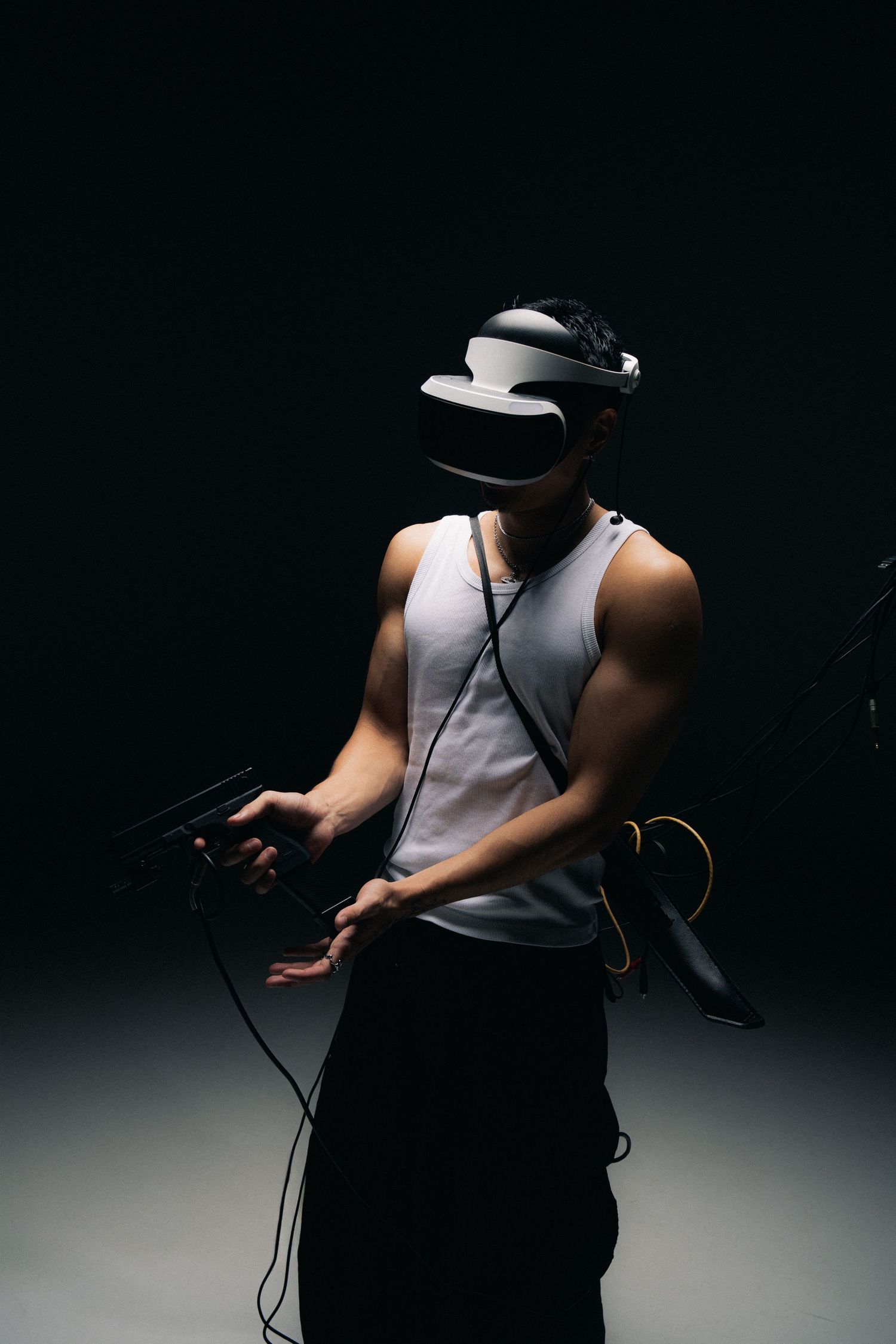Person wearing VR headset and tank top holding controllers in dramatic dark lighting against black background.