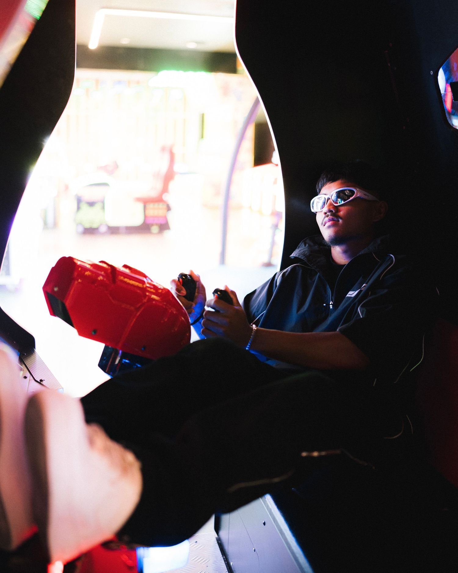 A person in dark clothing sits holding a red gaming controller in a dimly lit room.