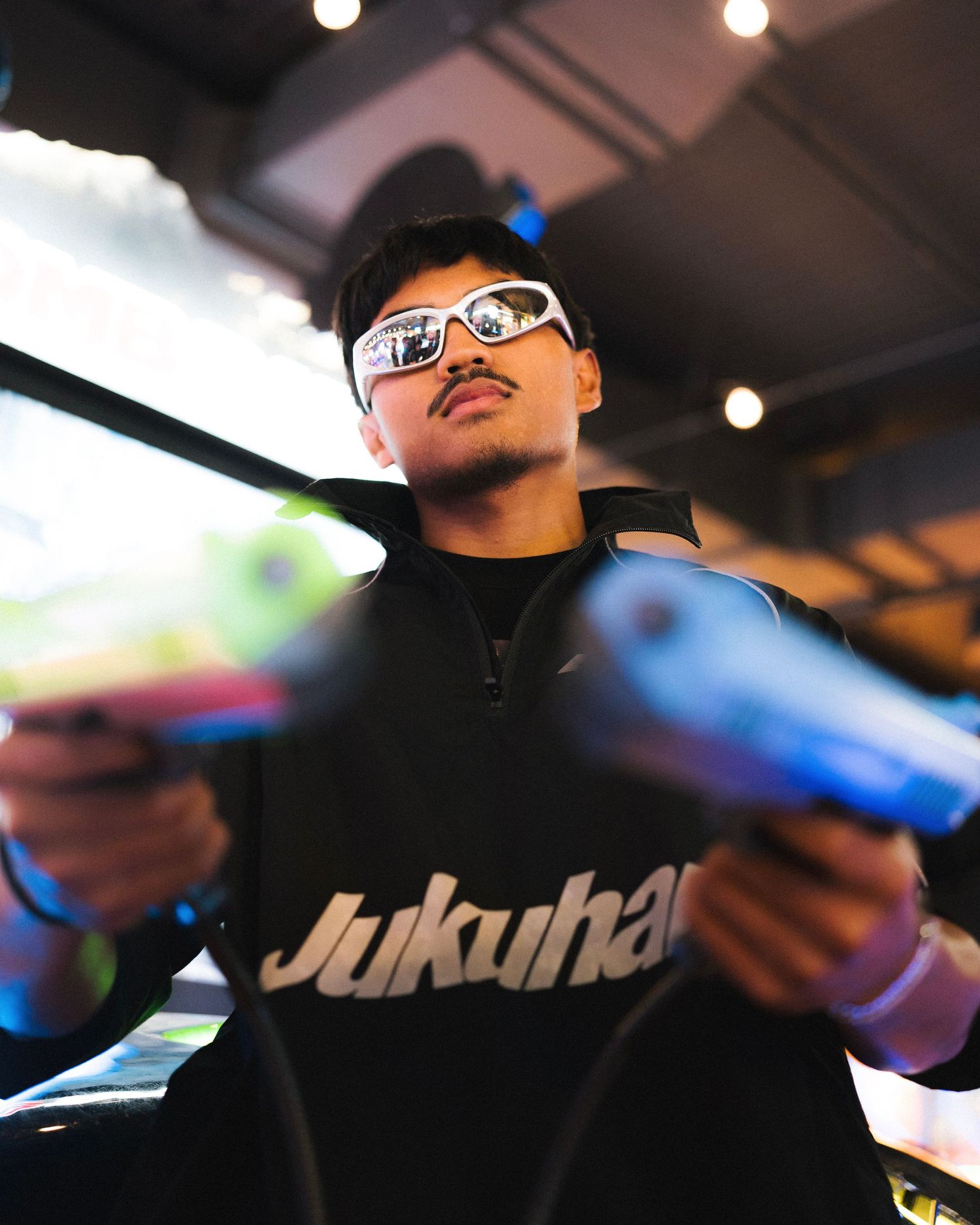 Someone wearing glasses and a dark Jukuhel sweatshirt holds blue items in a dimly lit indoor setting.