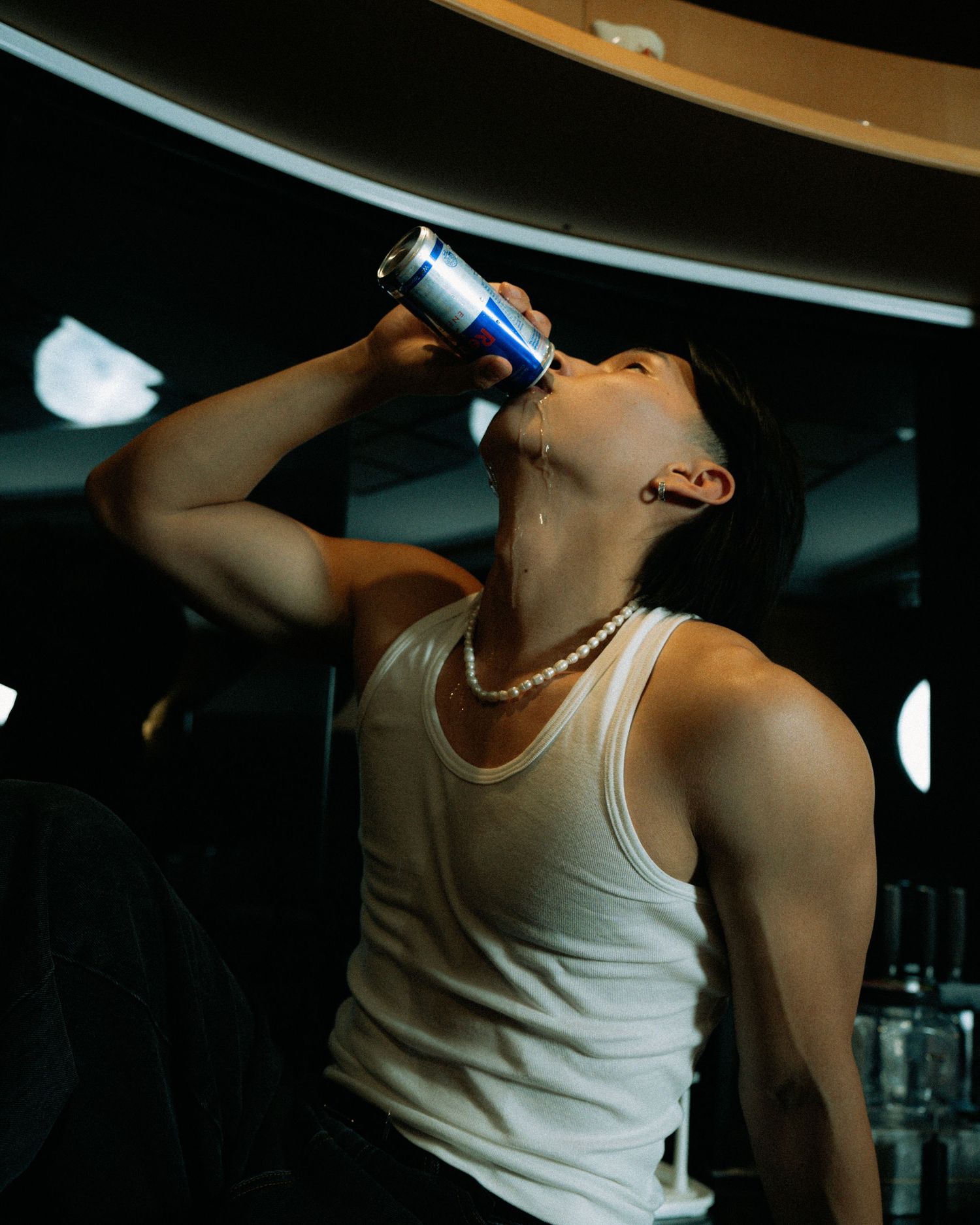 Person in white tank top drinks from a beverage can in dramatic dim lighting.