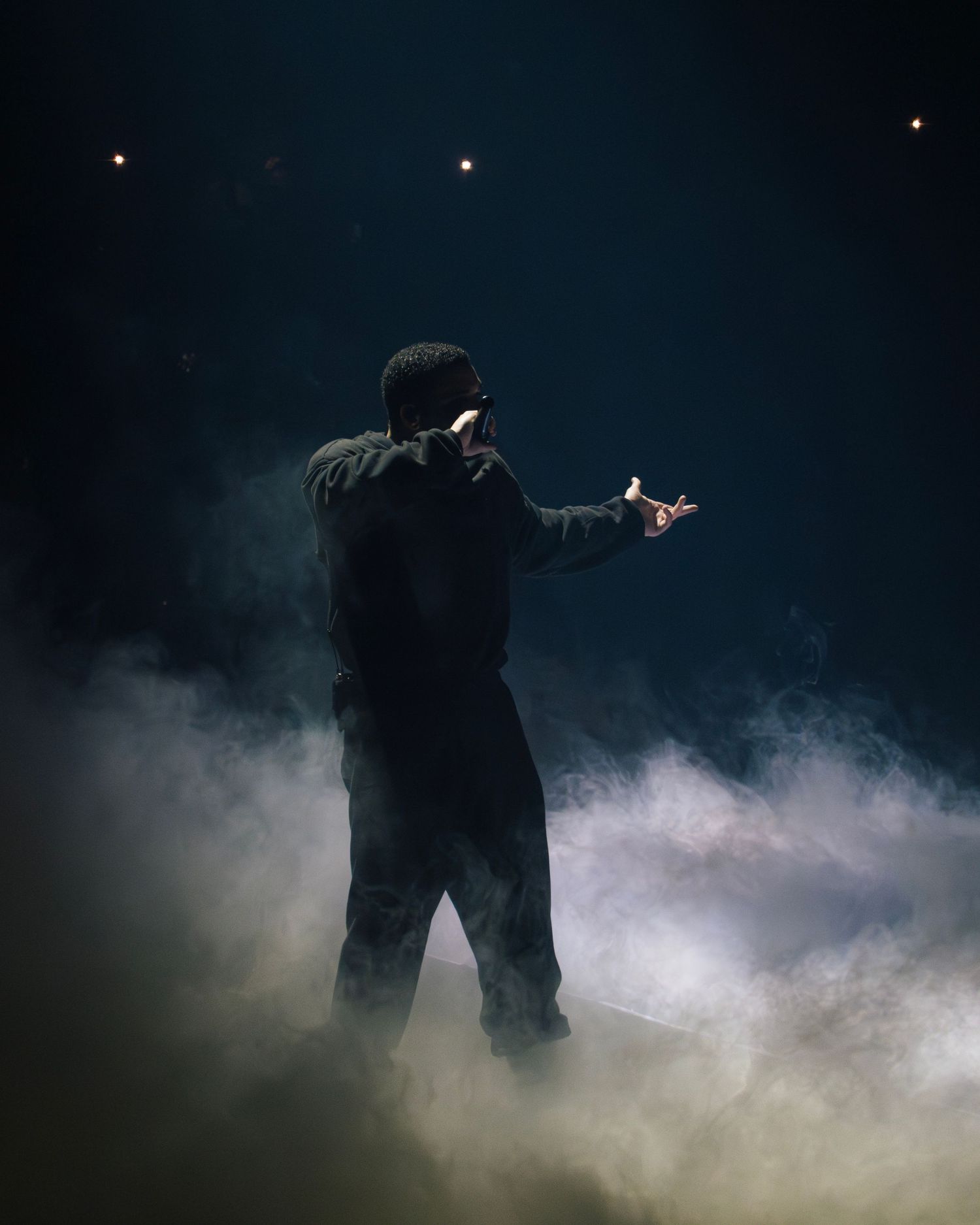 A silhouetted performer stands on stage amid dramatic fog effects and lighting in a dark atmosphere.