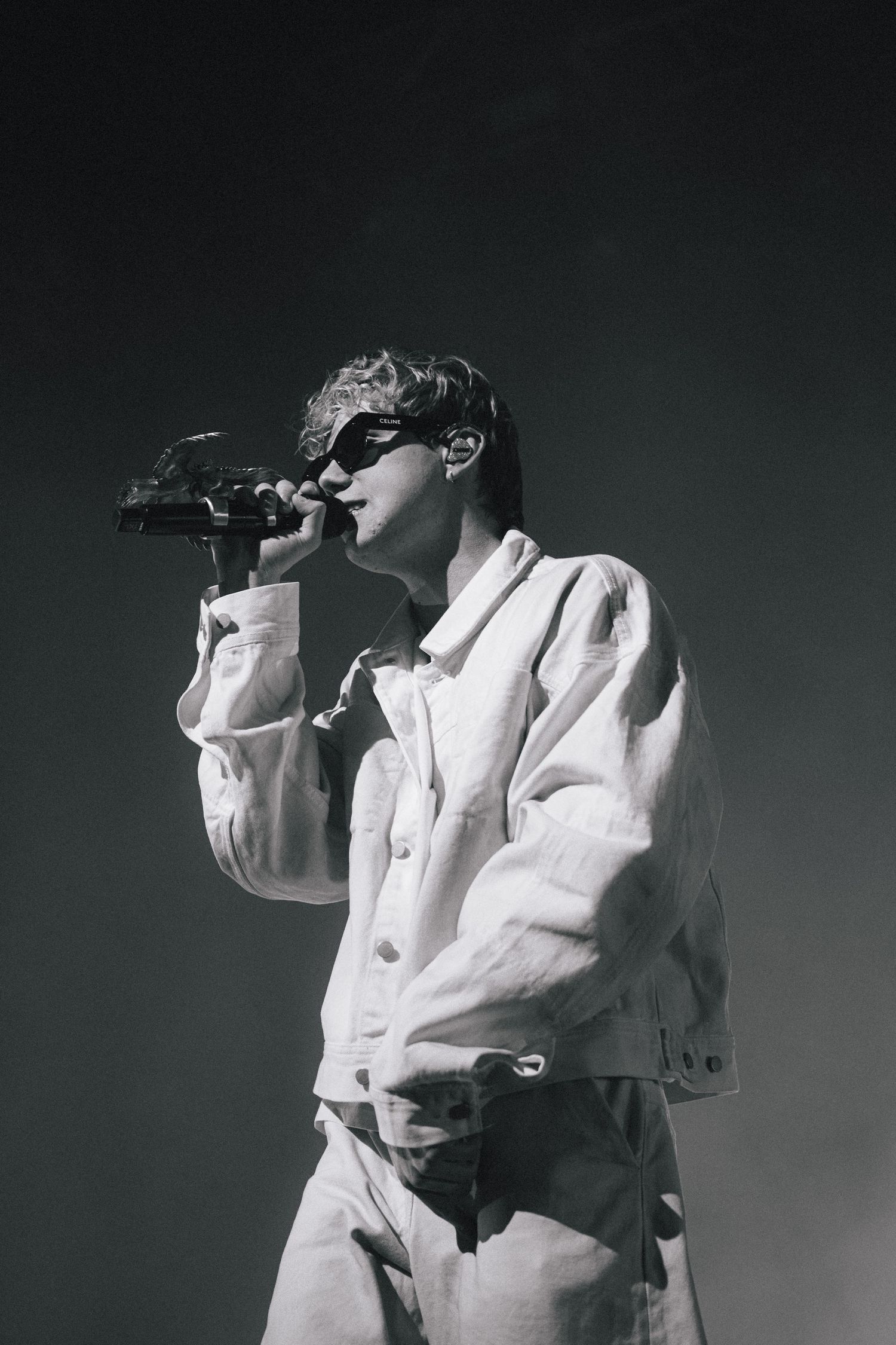 A performer in sunglasses and a white outfit sings into a microphone against a dark background in black and white.