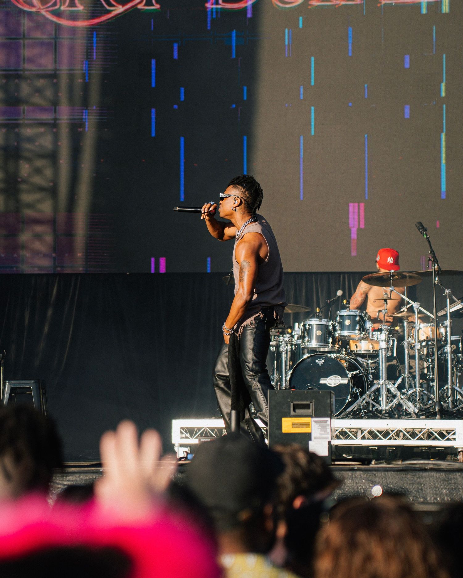 A performer sings passionately on stage during a concert with a drum kit visible in the background and colorful lighting effects.