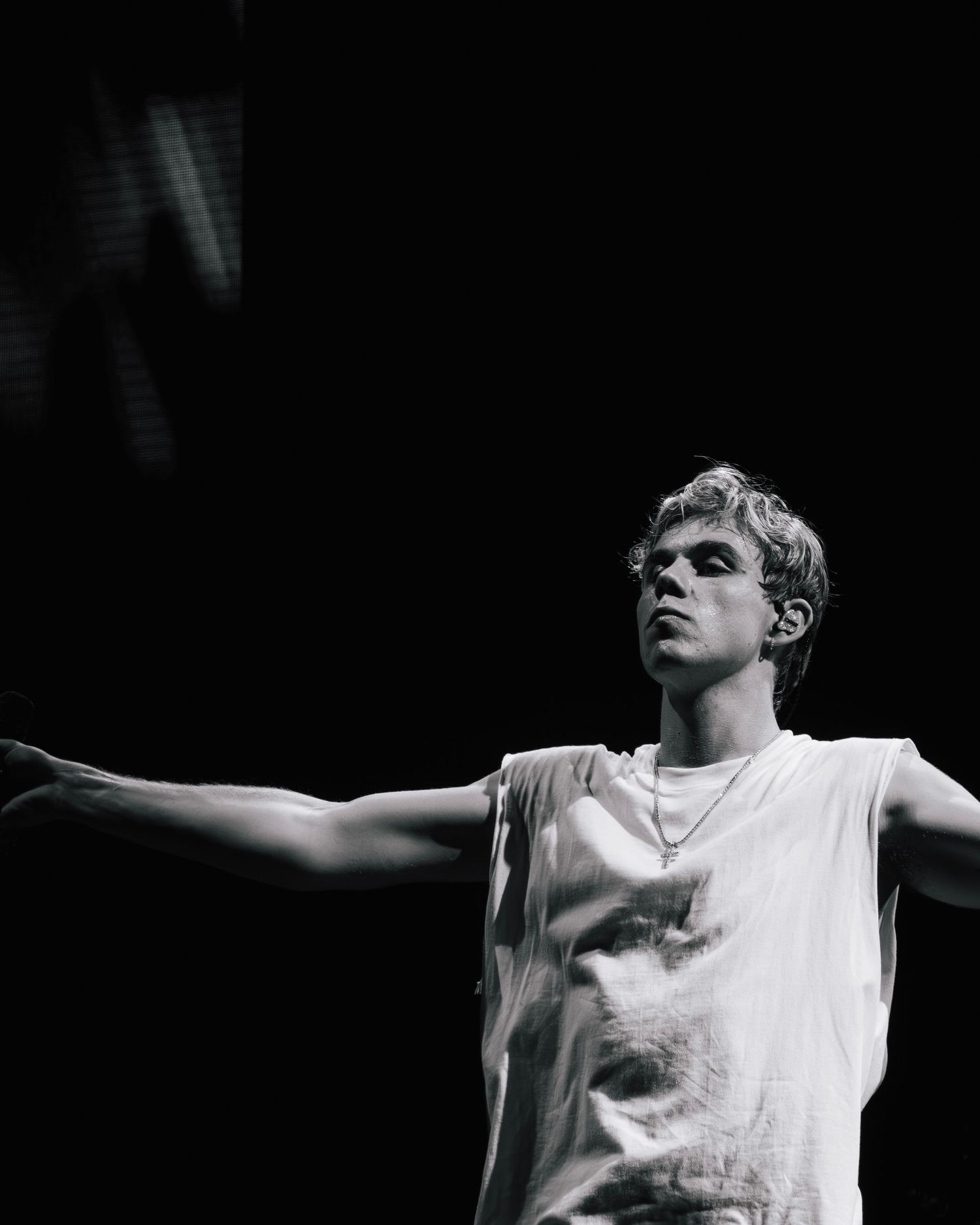 Black and white performance photo of a person in a white sleeveless shirt with arms outstretched against a dark background.