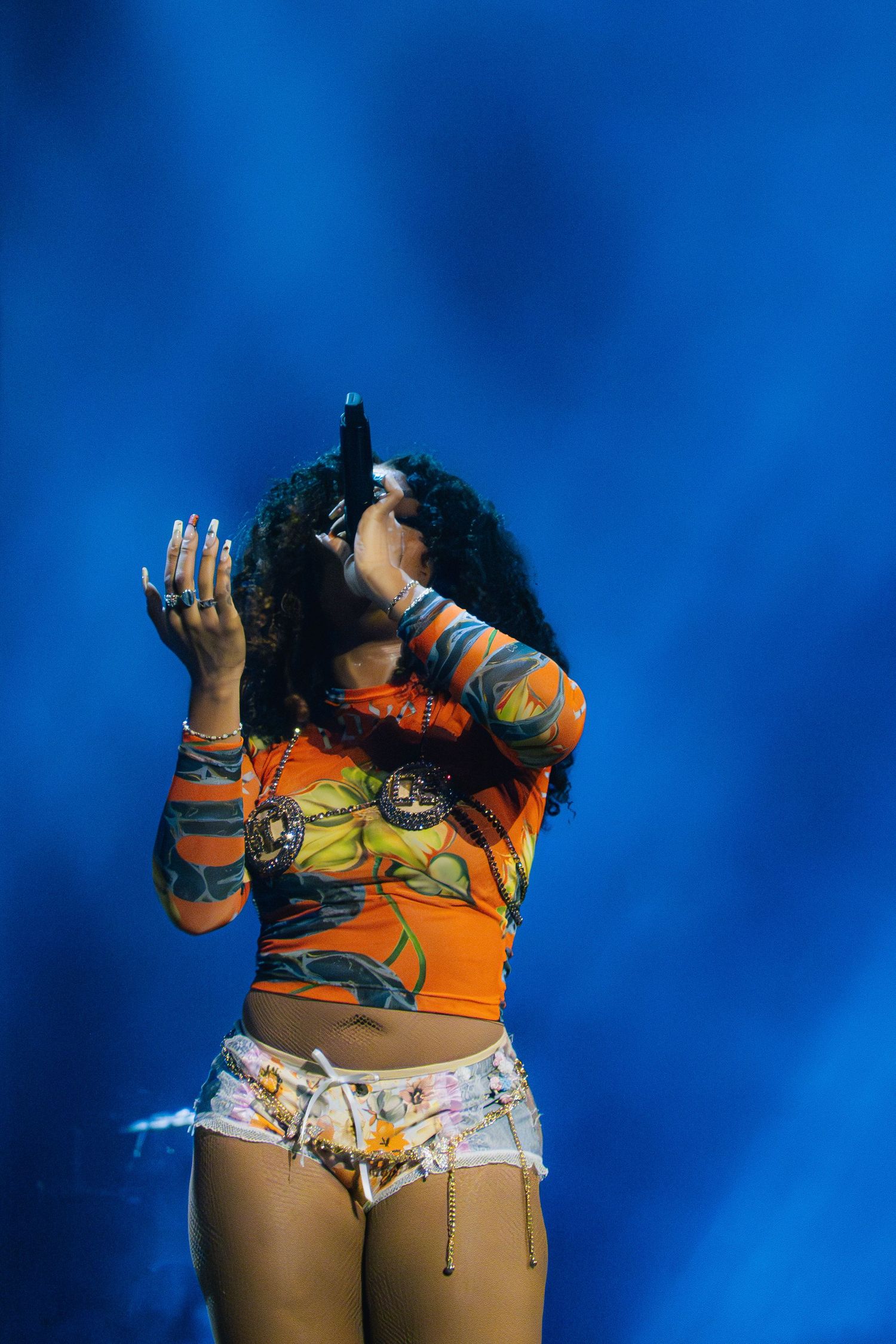 A performer in a colorful orange crop top and patterned shorts sings passionately against a vivid blue background.