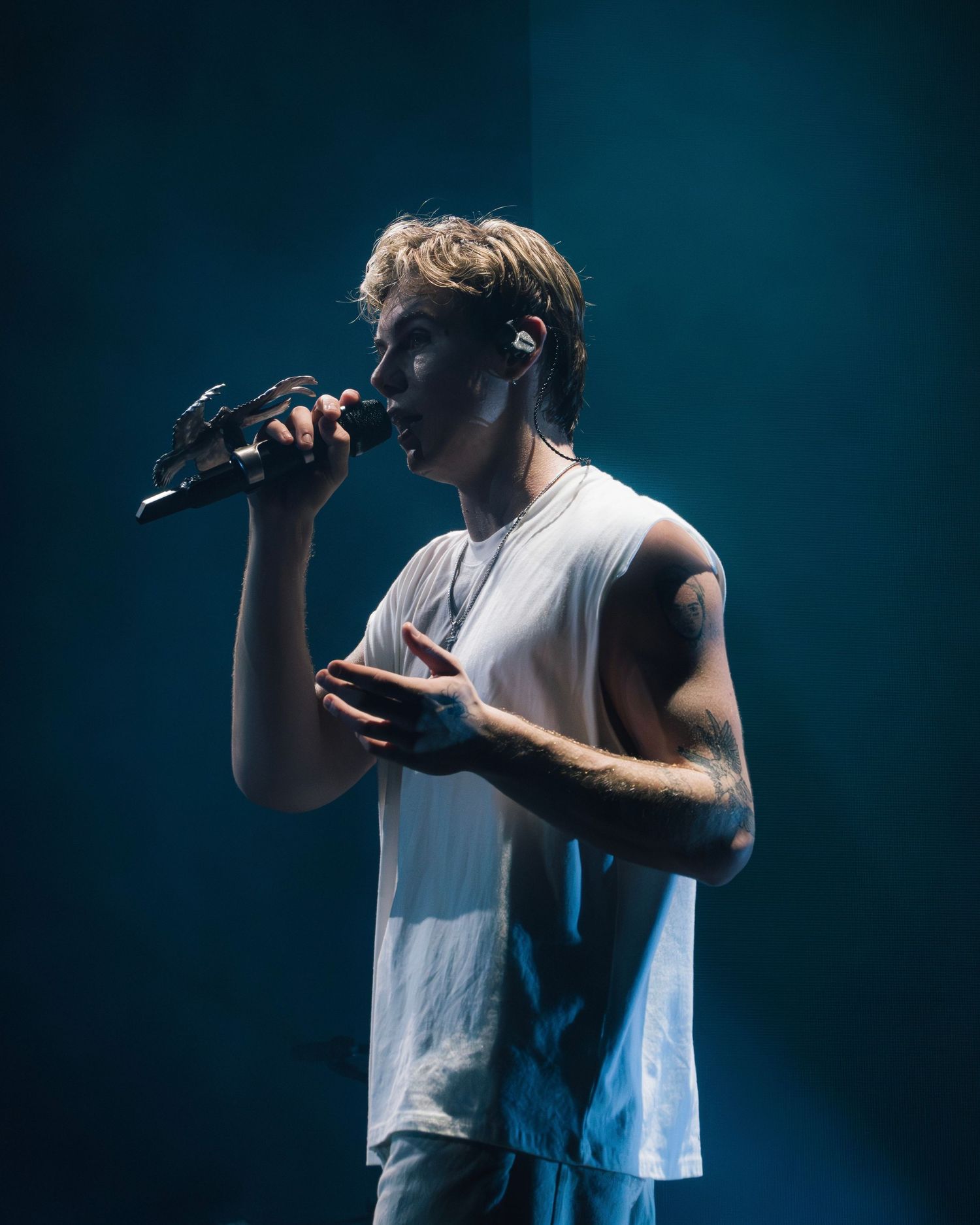 Singer performs on stage in a white tank top against a dark blue backdrop with dramatic lighting effects.
