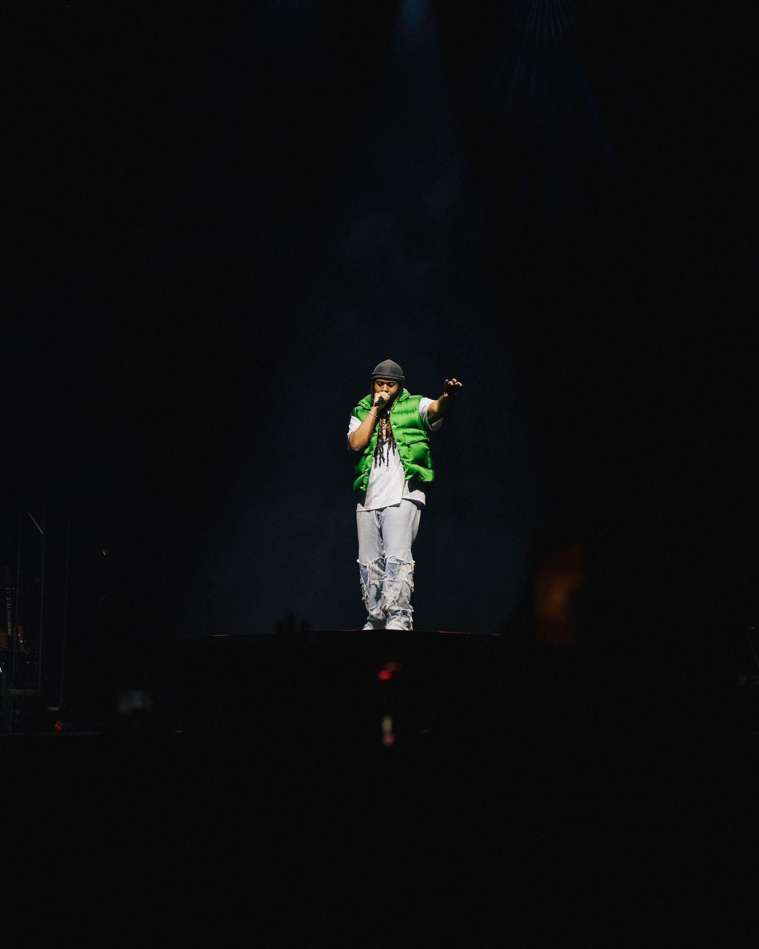 A performer in a green jacket and light pants stands illuminated by a spotlight on a dark stage.