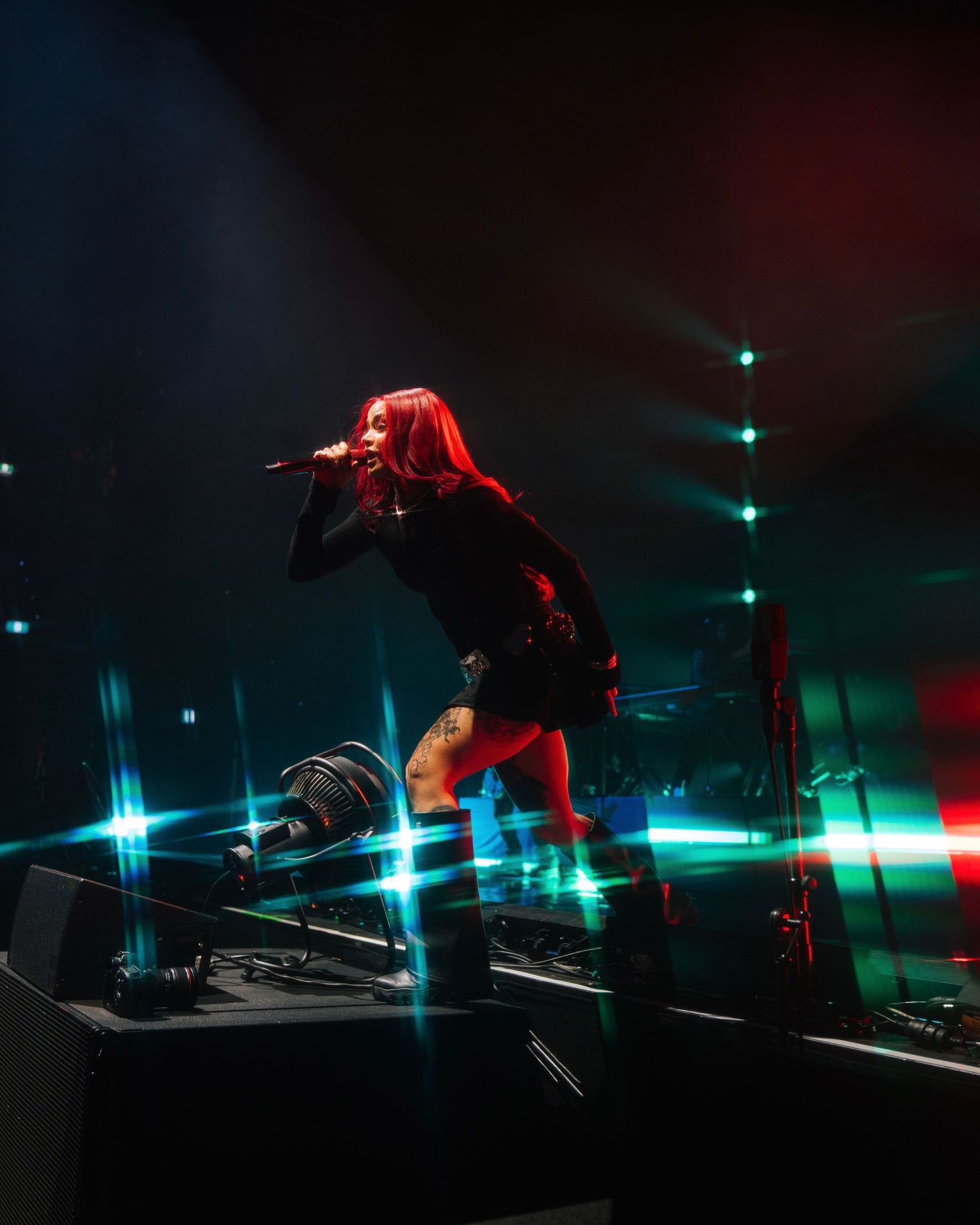 Energetic performer with red hair leaps on stage amidst dramatic lighting with green and red tones during concert performance.