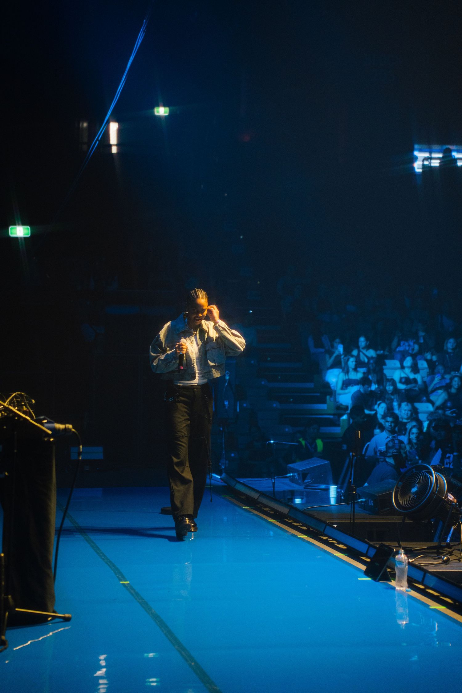 A performer sings on stage illuminated by blue lighting during a concert with an audience visible in the background.