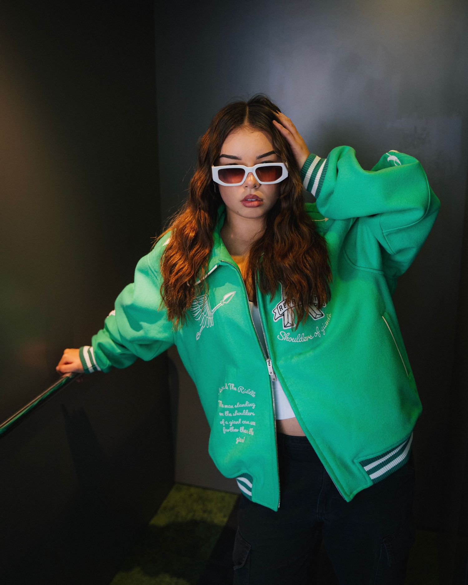 Person in green varsity jacket and white sunglasses poses against dark background.