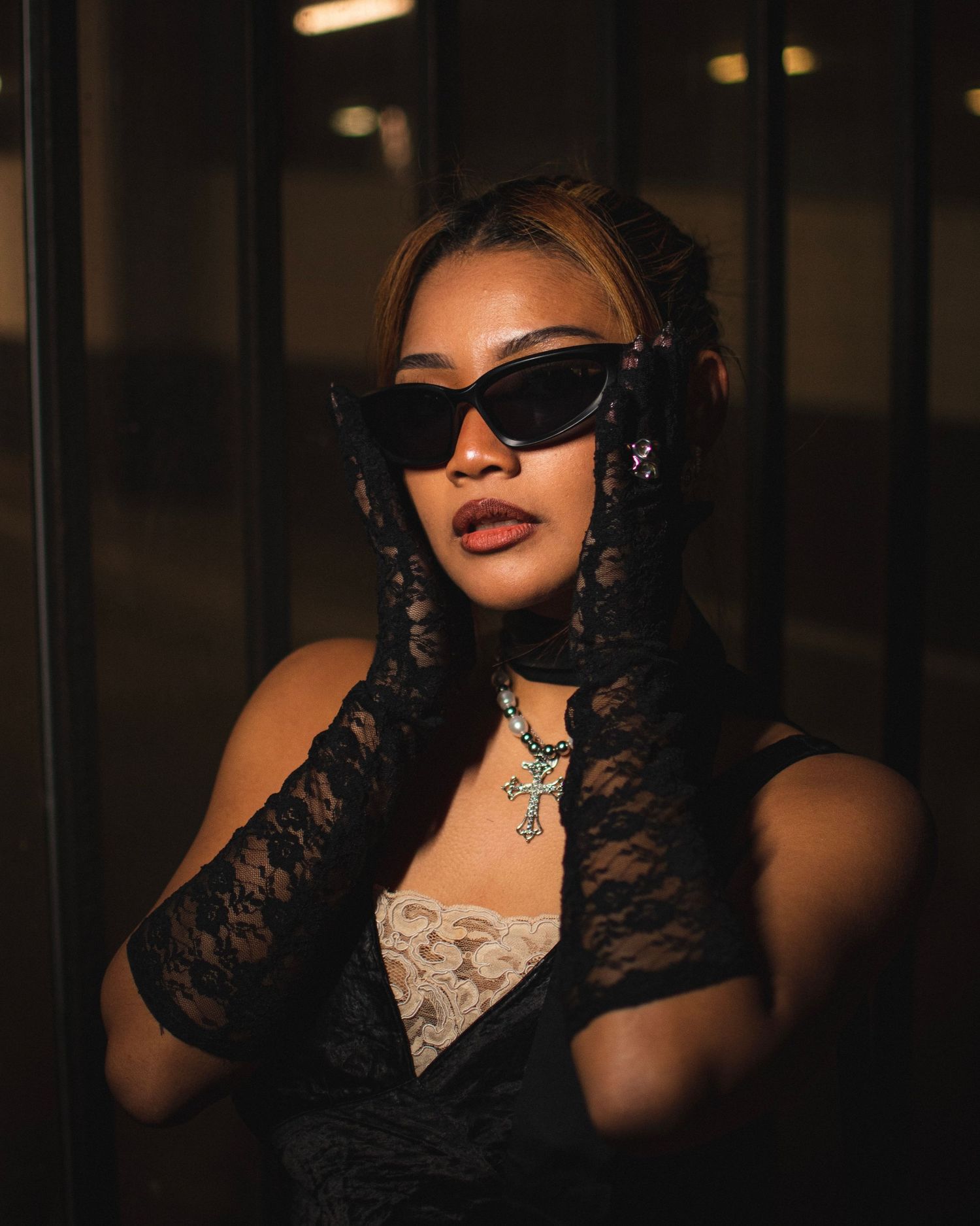 Glamorous portrait featuring sunglasses, lace gloves and statement jewelry in dim lighting.