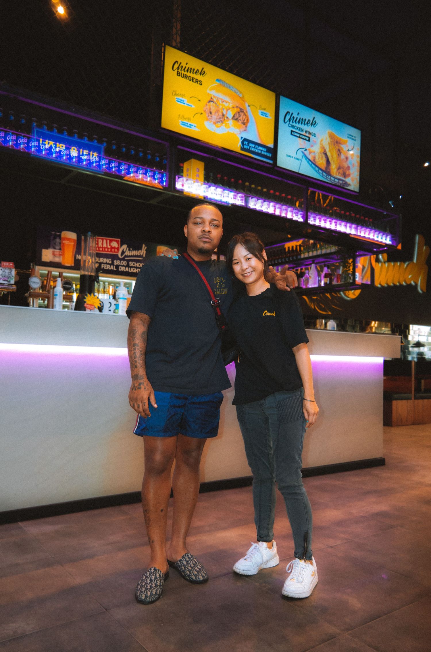 Casual portrait of two people standing together in front of illuminated bar counter.