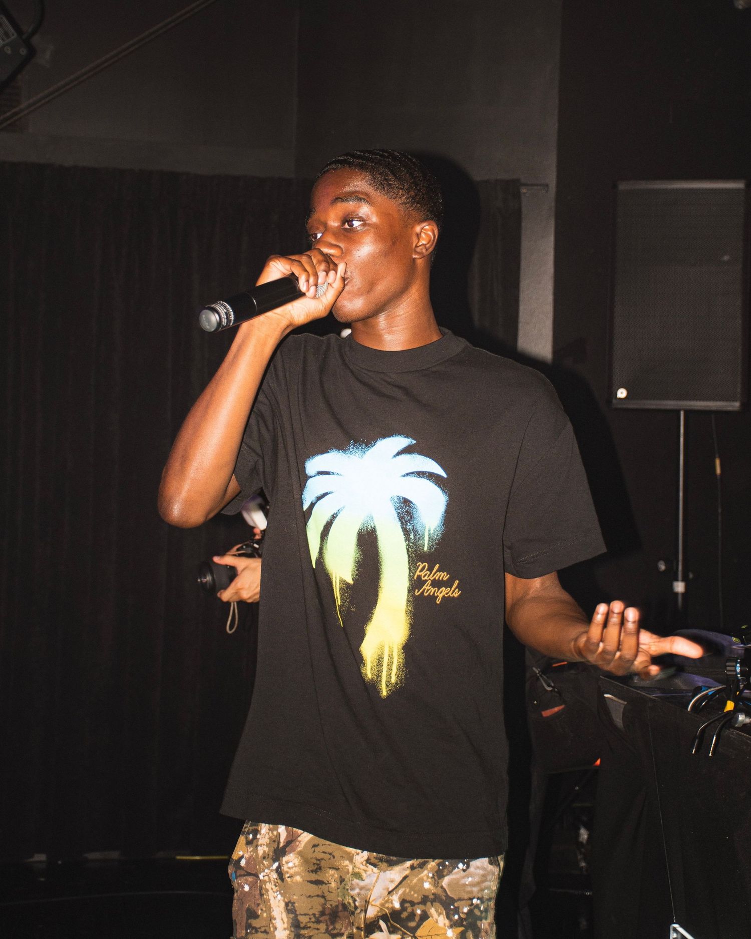 Performer wearing palm tree graphic t-shirt singing into microphone on dark stage.