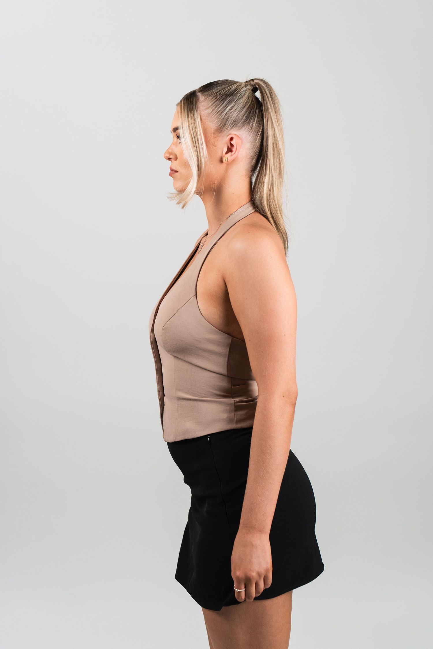 Side profile view of a person in a beige sleeveless top and black skirt posing against a light gray background.