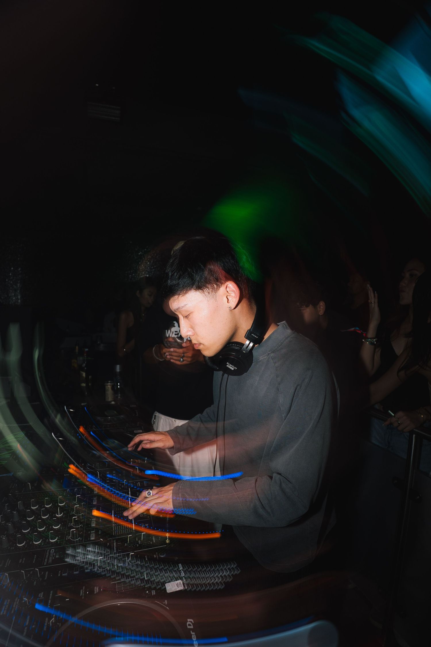 A DJ performs at a nightclub with headphones and equipment under colorful green and blue lighting.