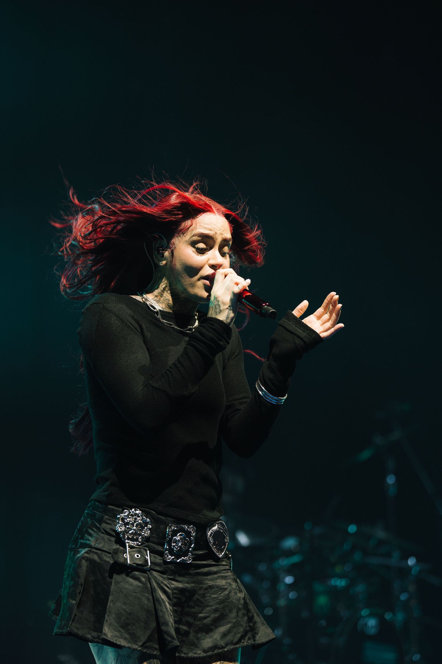A performer with red flowing hair singing passionately on stage in a black outfit with a decorative belt.