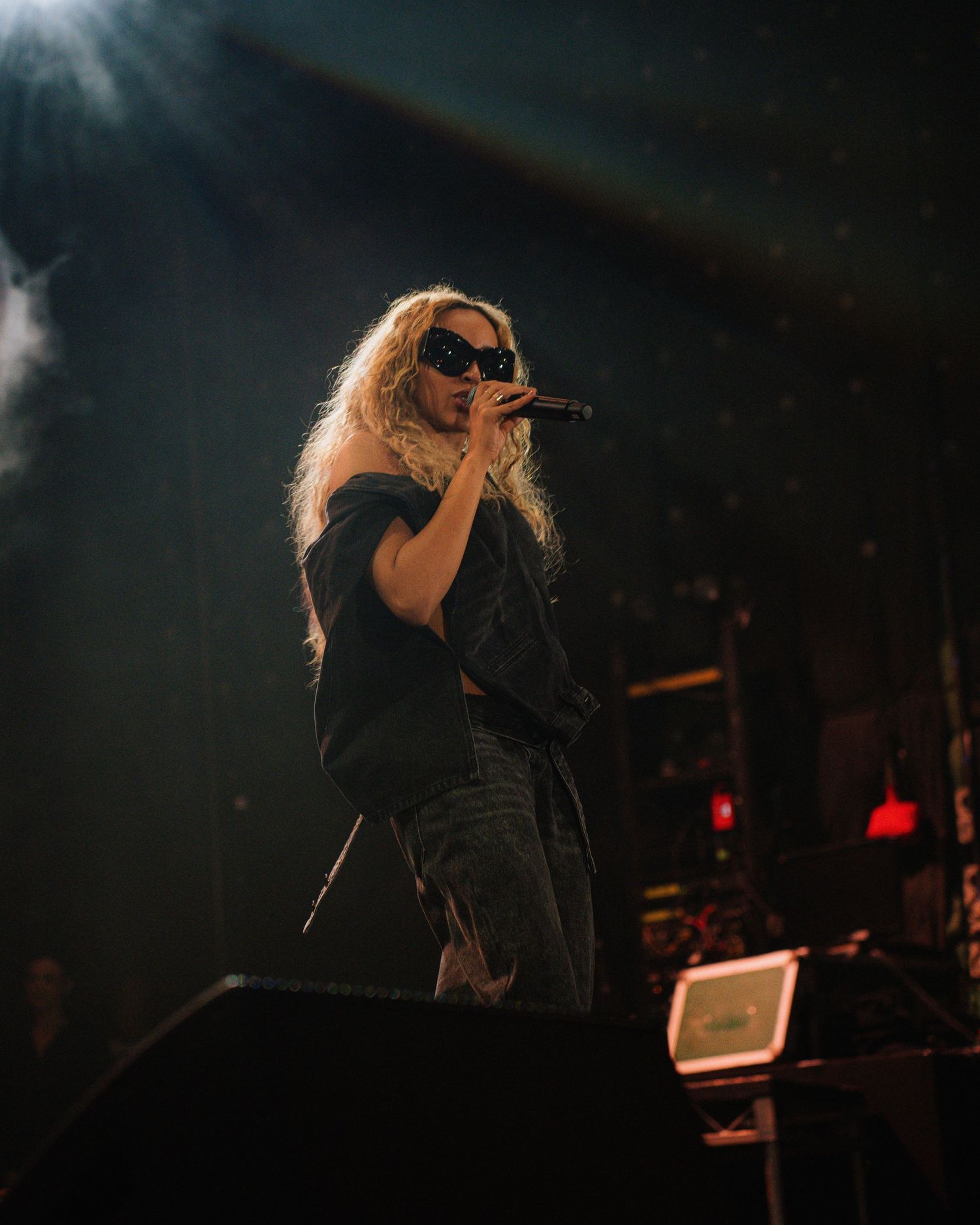 Concert performer with long hair in black outfit singing on dark stage with dramatic lighting.