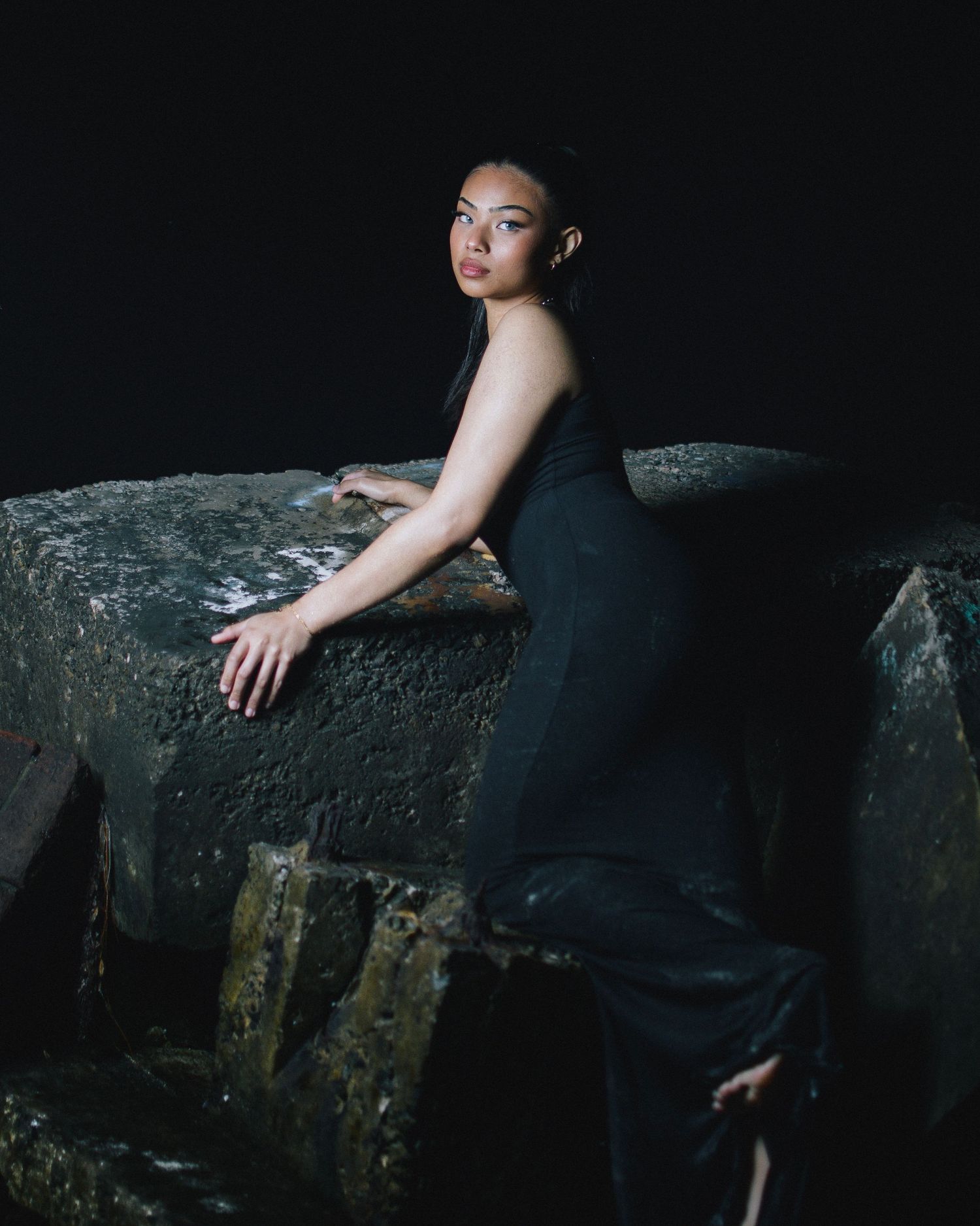 A person in a black evening dress poses dramatically against a dark textured wall in moody lighting.