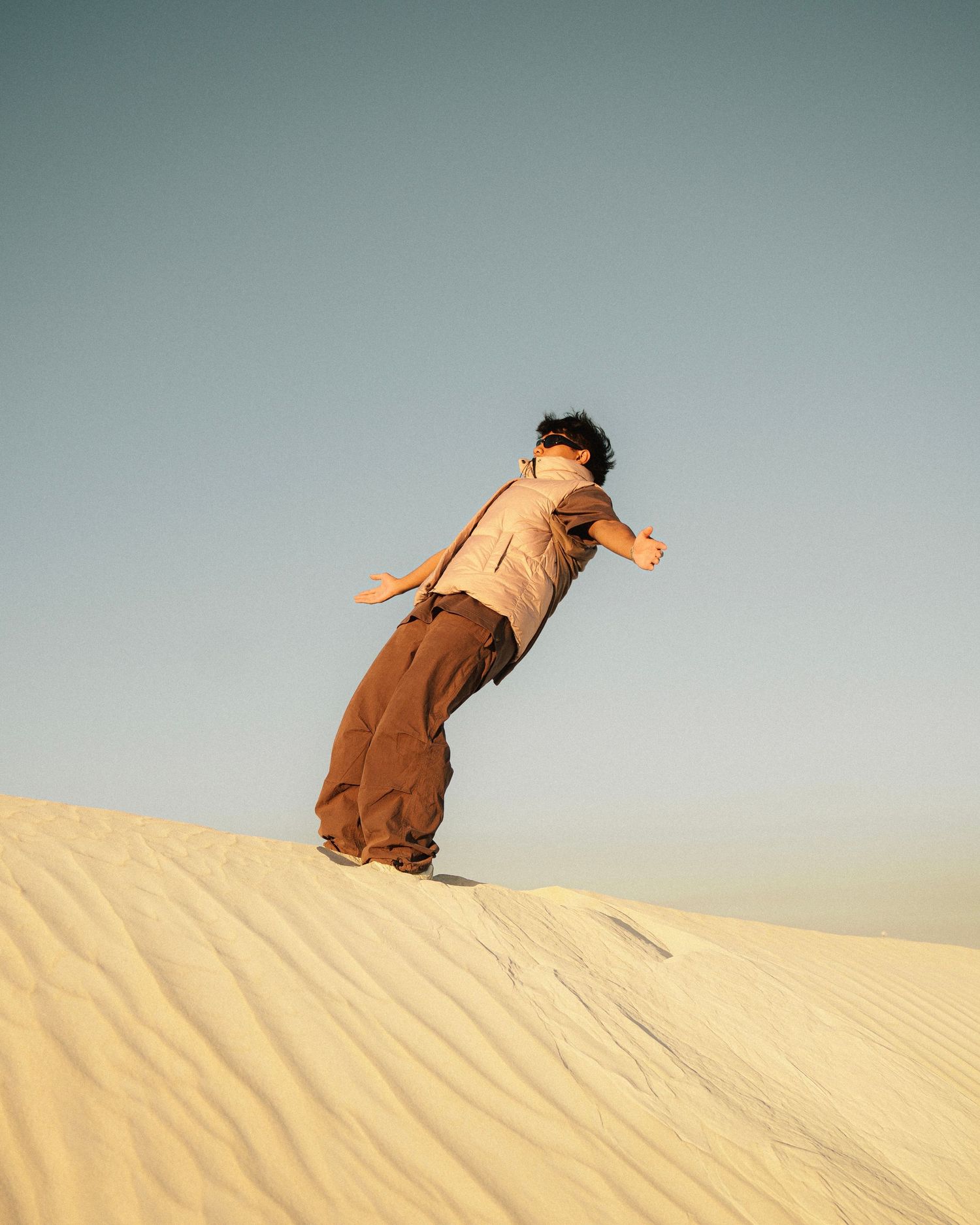 Person leaning back dramatically on desert sand dune against blue sky.