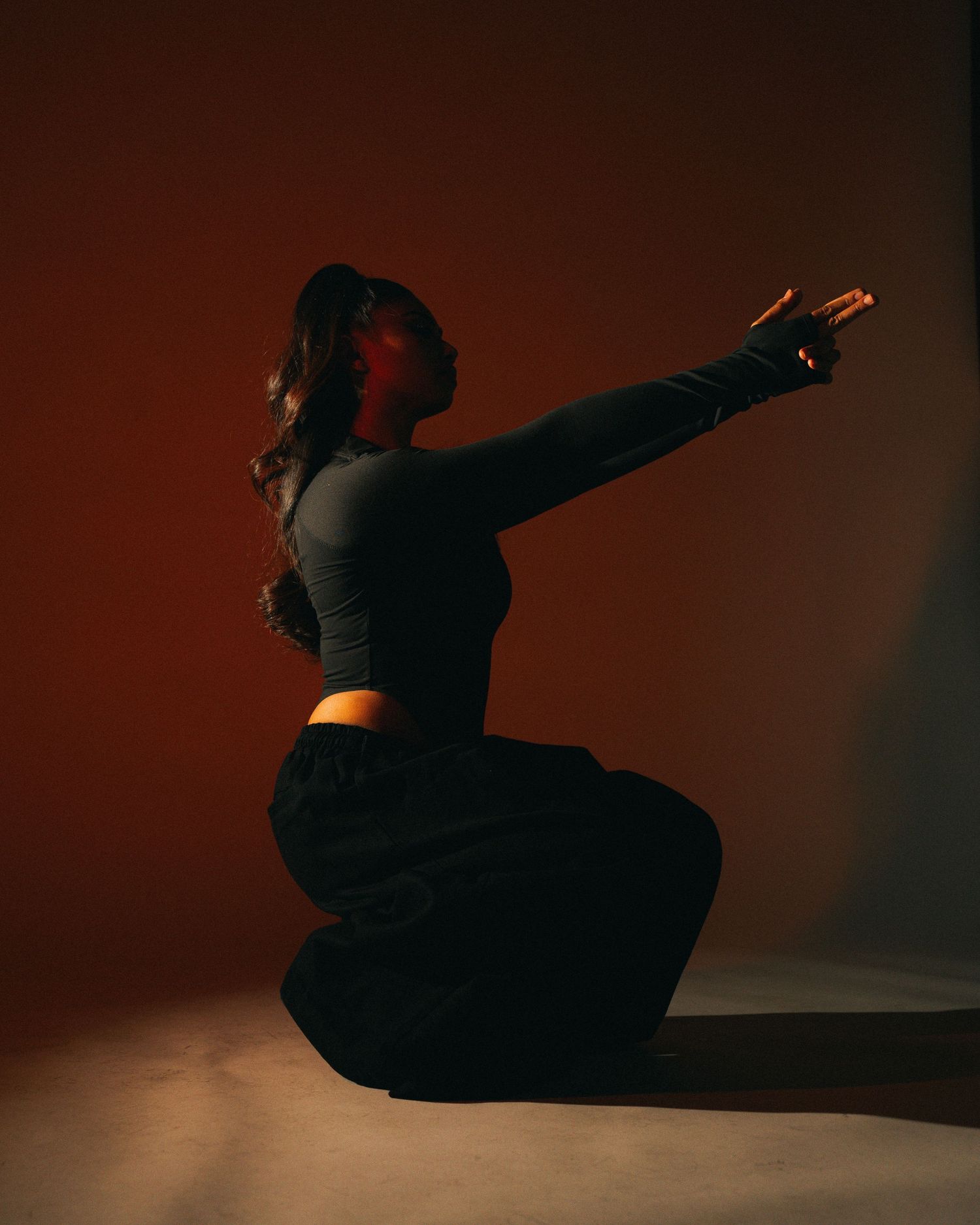 Figure in black clothing performs dramatic pose against dark red background in artistic portrait.